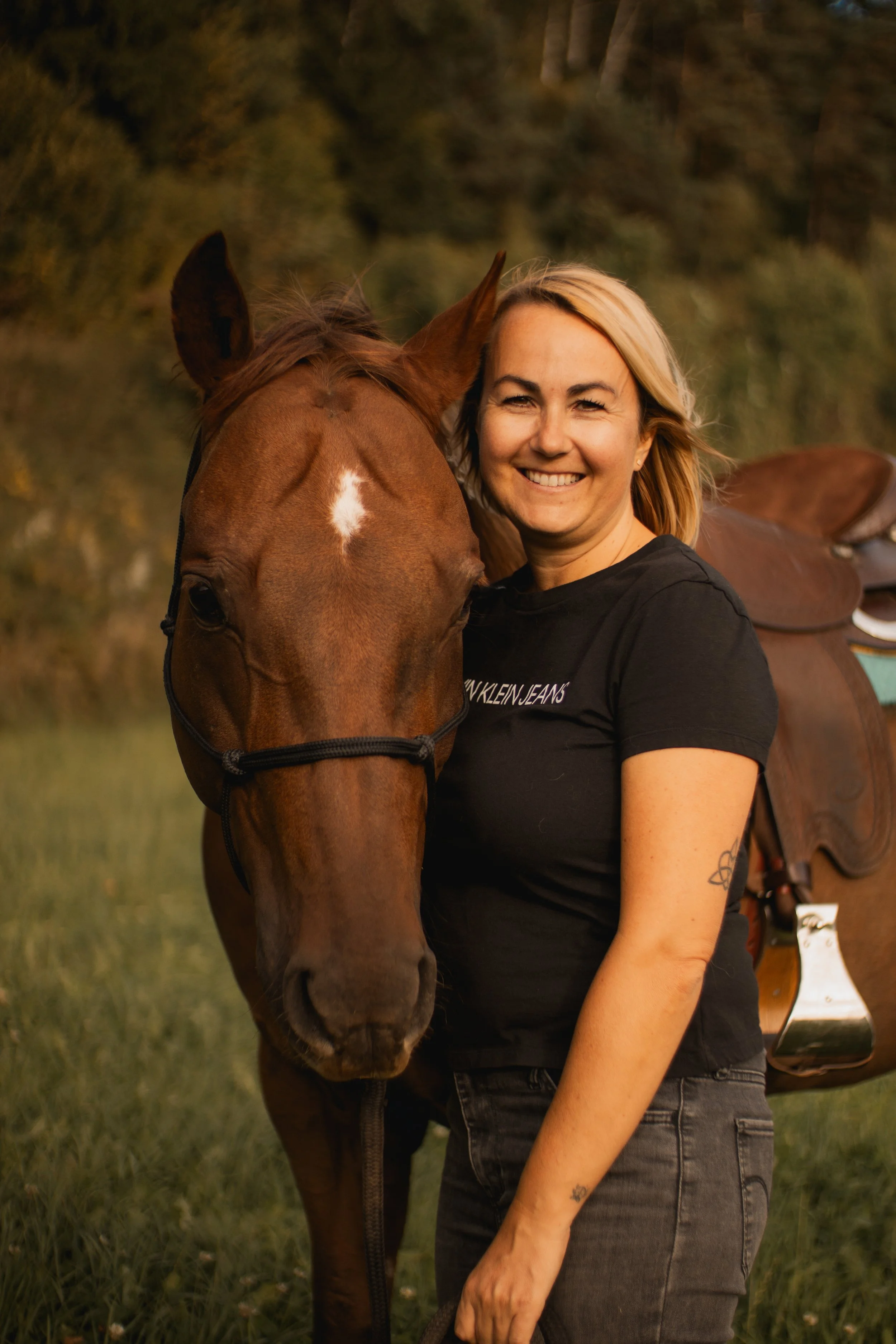 Eine lachende Frau mit kurzen, blonden Haaren steht neben einem braunen Pferd mit Sattel im Freien, umgeben von Bäumen und Gras bei Sonnenuntergang.