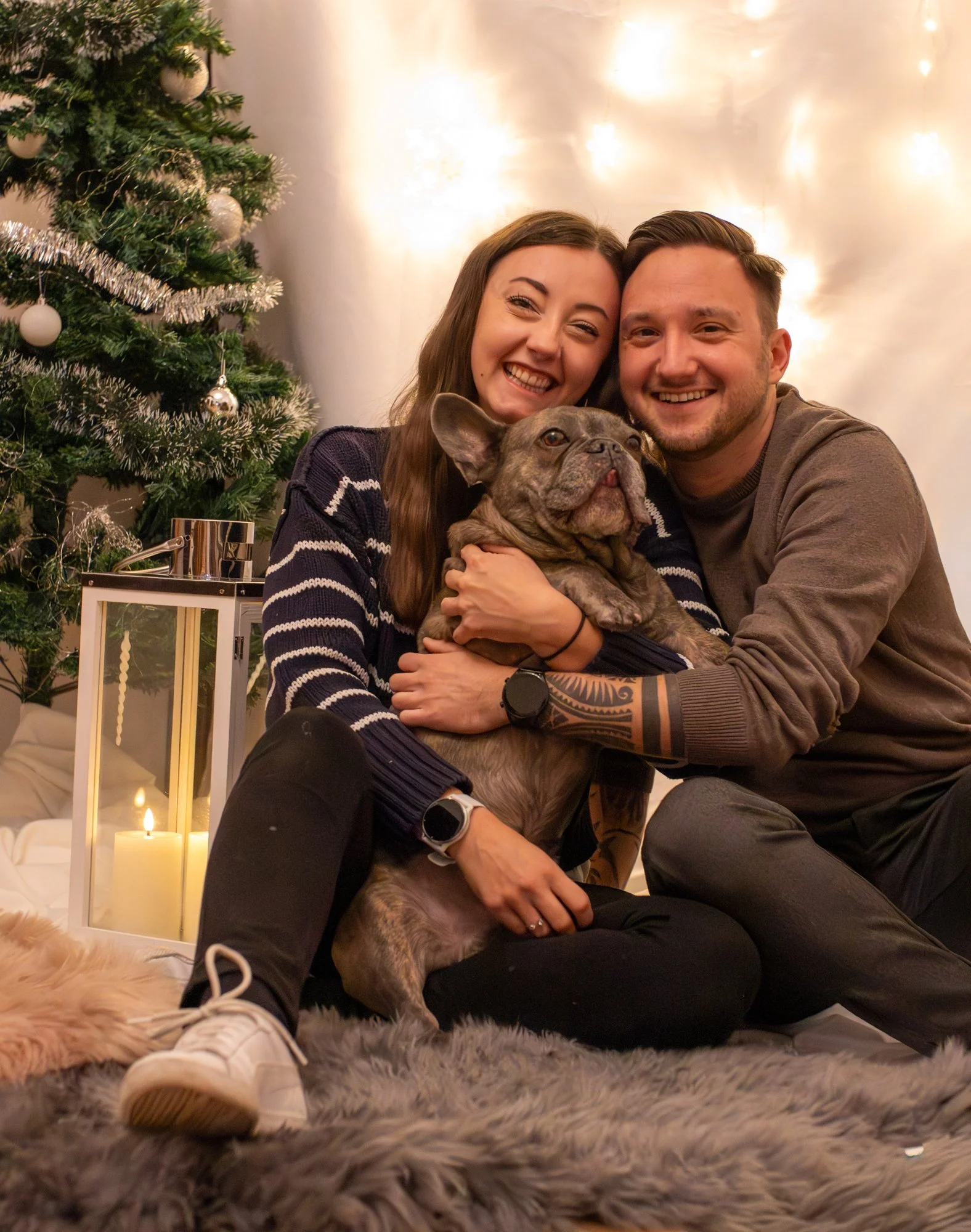 Ein lachendes junges Paar mit einem französischen Bulldoggen-Hund vor einem festlich geschmückten Weihnachtsbaum, umgeben von Kerzen und weichem Teppich.