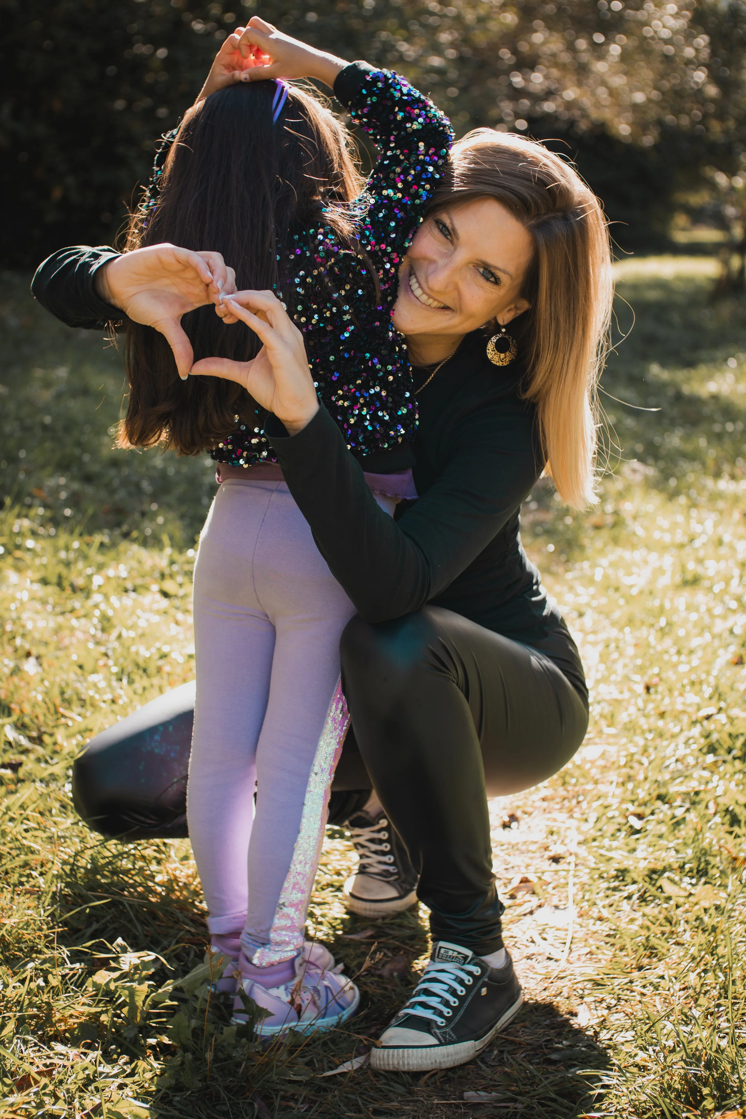 Eine Frau umarmt ein Mädchen im Freien, beide lächeln und machen Herzhandzeichen, umgeben von Natur bei Sonnenuntergang.