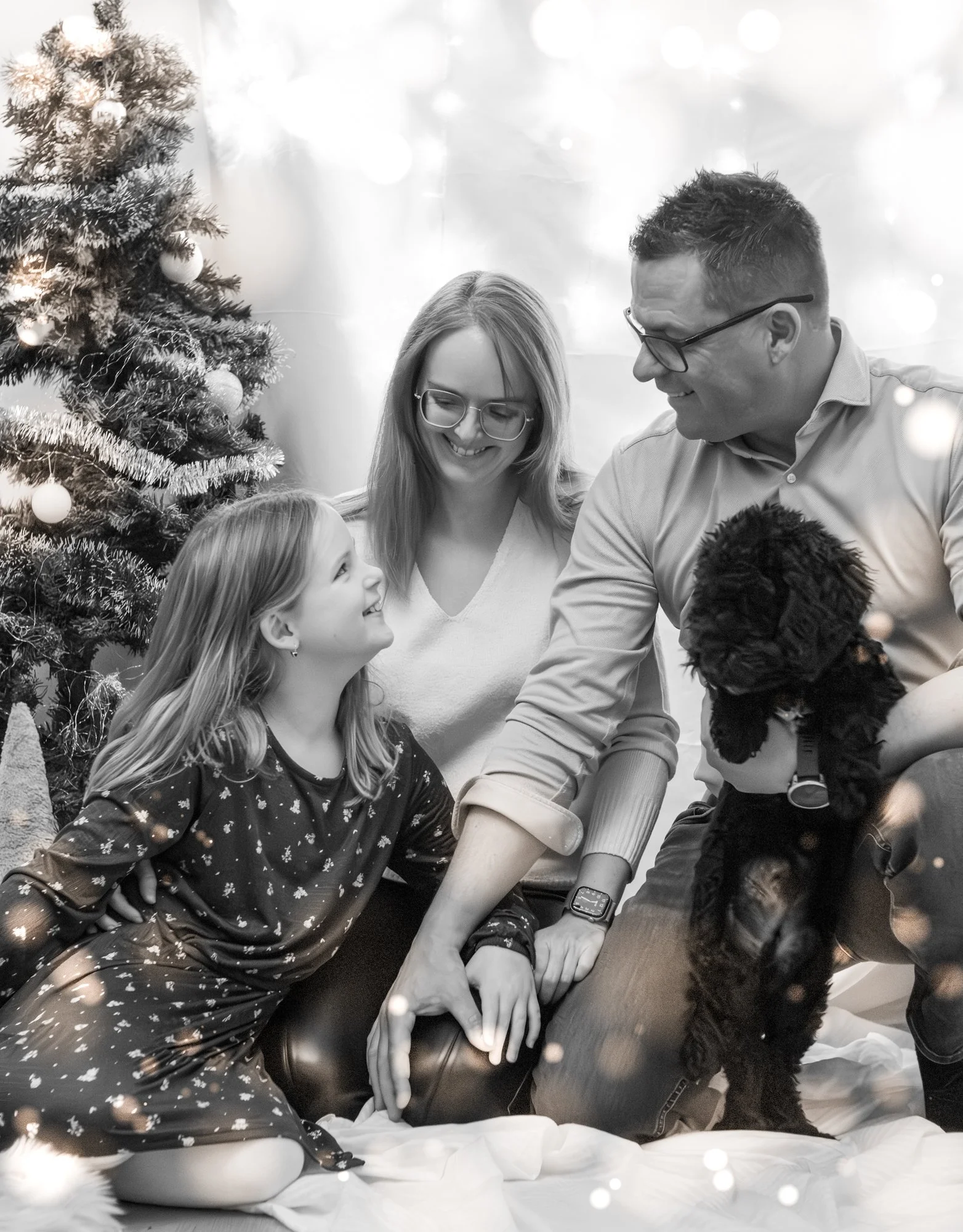 Familie mit Kind und Hund bei Weihnachtsfeier vor Weihnachtsbaum, fröhlich lachend, schwarz-weiß Foto.