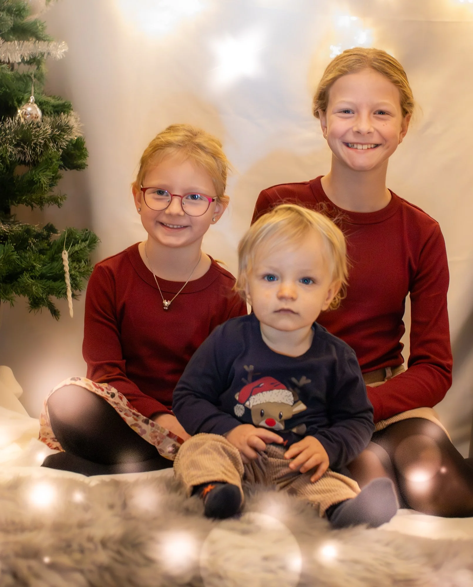 Drei Kinder sitzen auf einem weißen Fellteppich vor einem Weihnachtsbaum, der mit silbernen Kugeln dekoriert ist. Es ist Weihnachten, und die Kinder tragen festliche Kleidung.