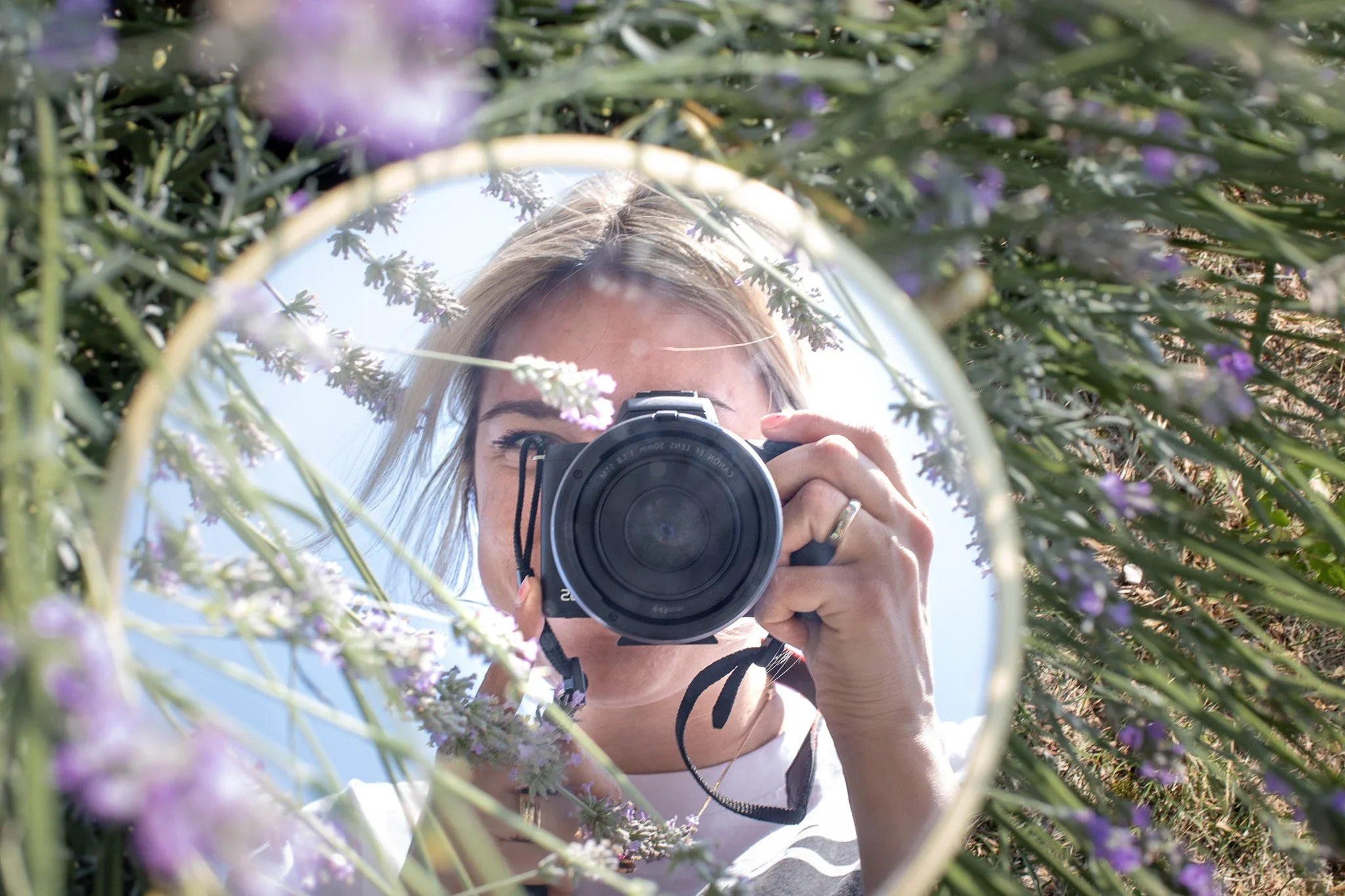 Eine Person fotografiert sich selbst mit einer Kamera in einem runden Spiegel, umgeben von grünen Pflanzen und lila Blumen im Freien.