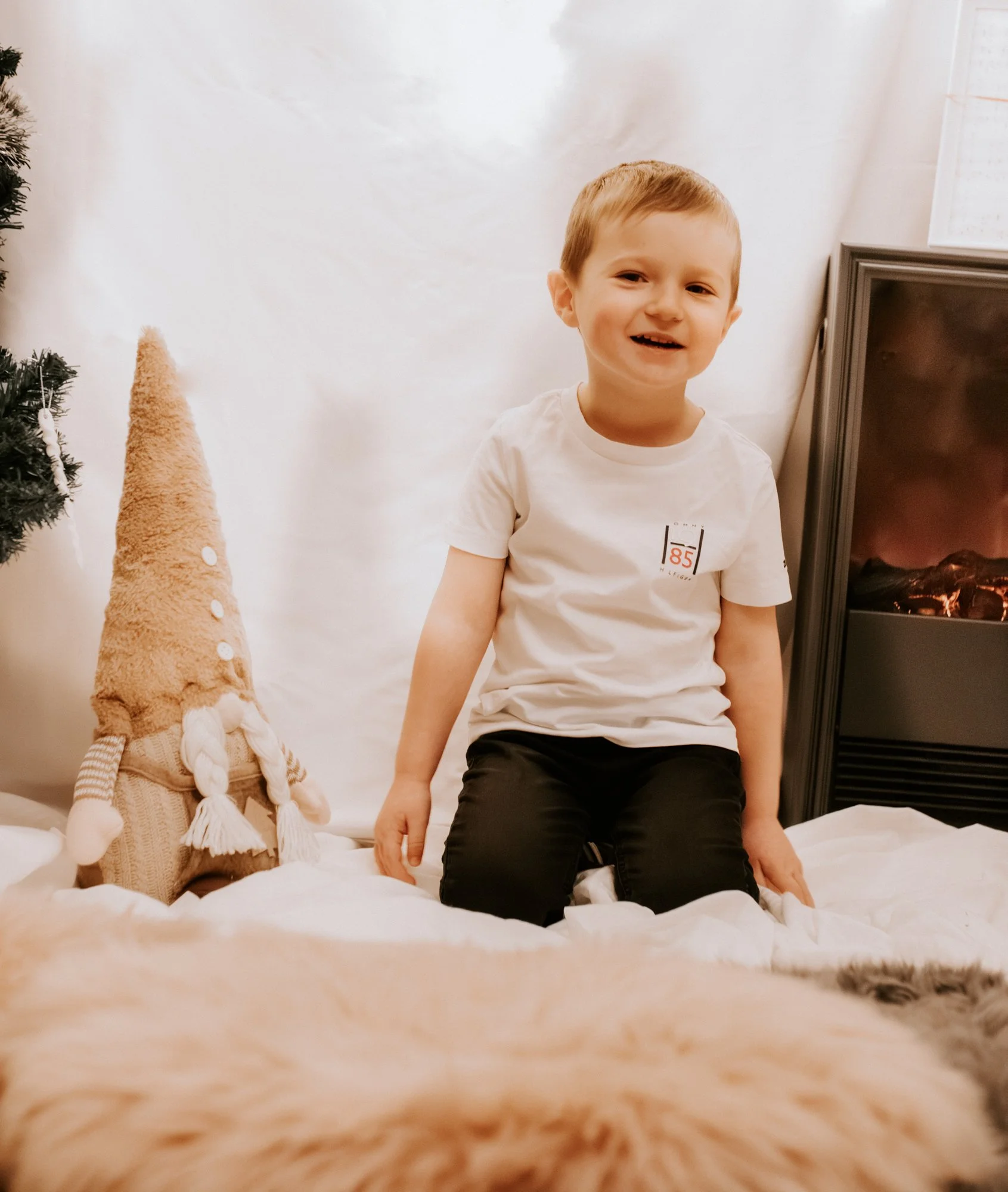 Ein Junge sitzt auf einem weißen Bett vor einem Kamin und einem Weihnachtsbaum. Neben ihm steht eine dekorative Weihnachtsfigur in Form eines Gnom. Im Vordergrund ist das Fell eines Tieres zu sehen.