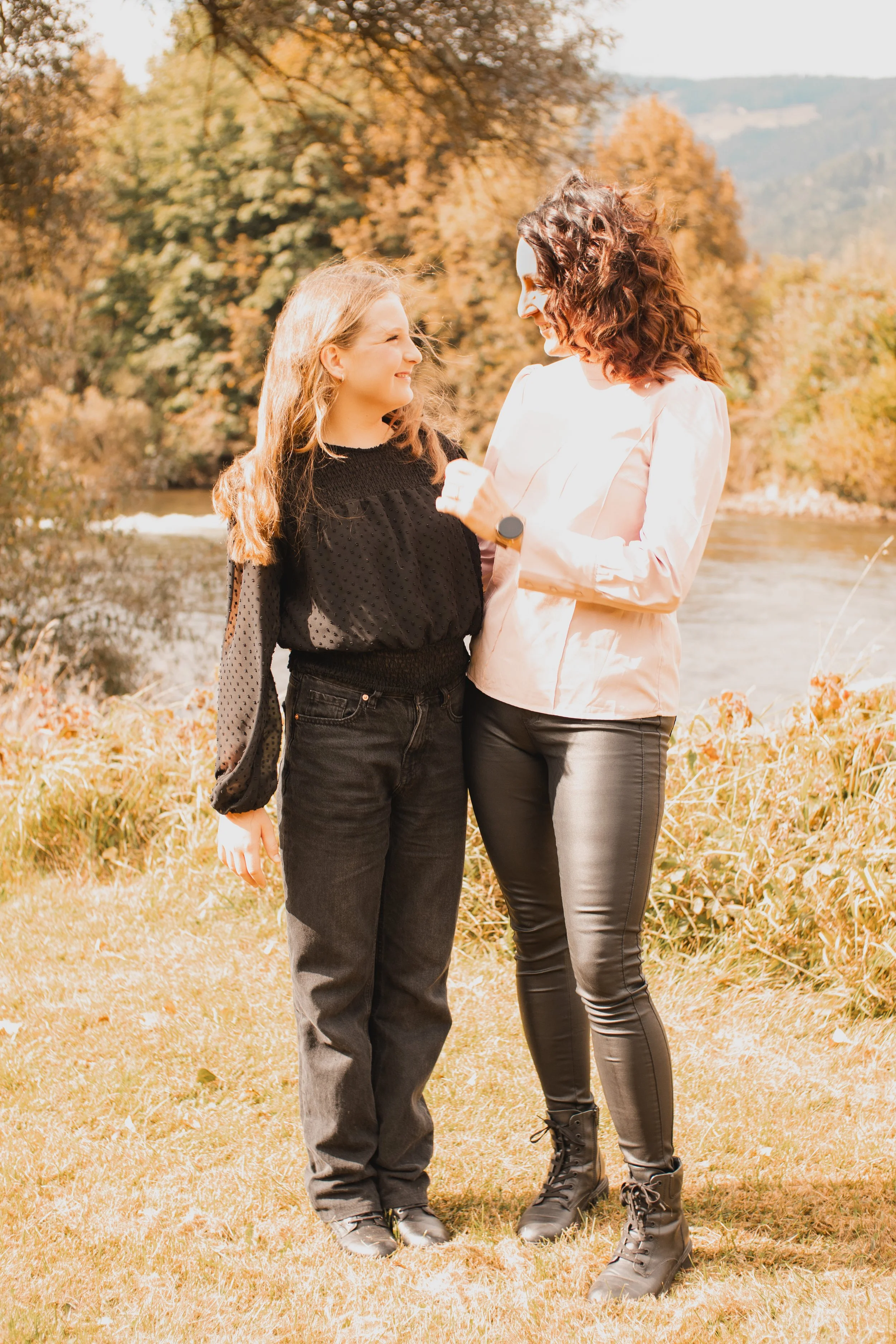 Zwei Frauen stehen sich lachend am Flussufer mit herbstlicher Baumlandschaft im Hintergrund gegenüber.