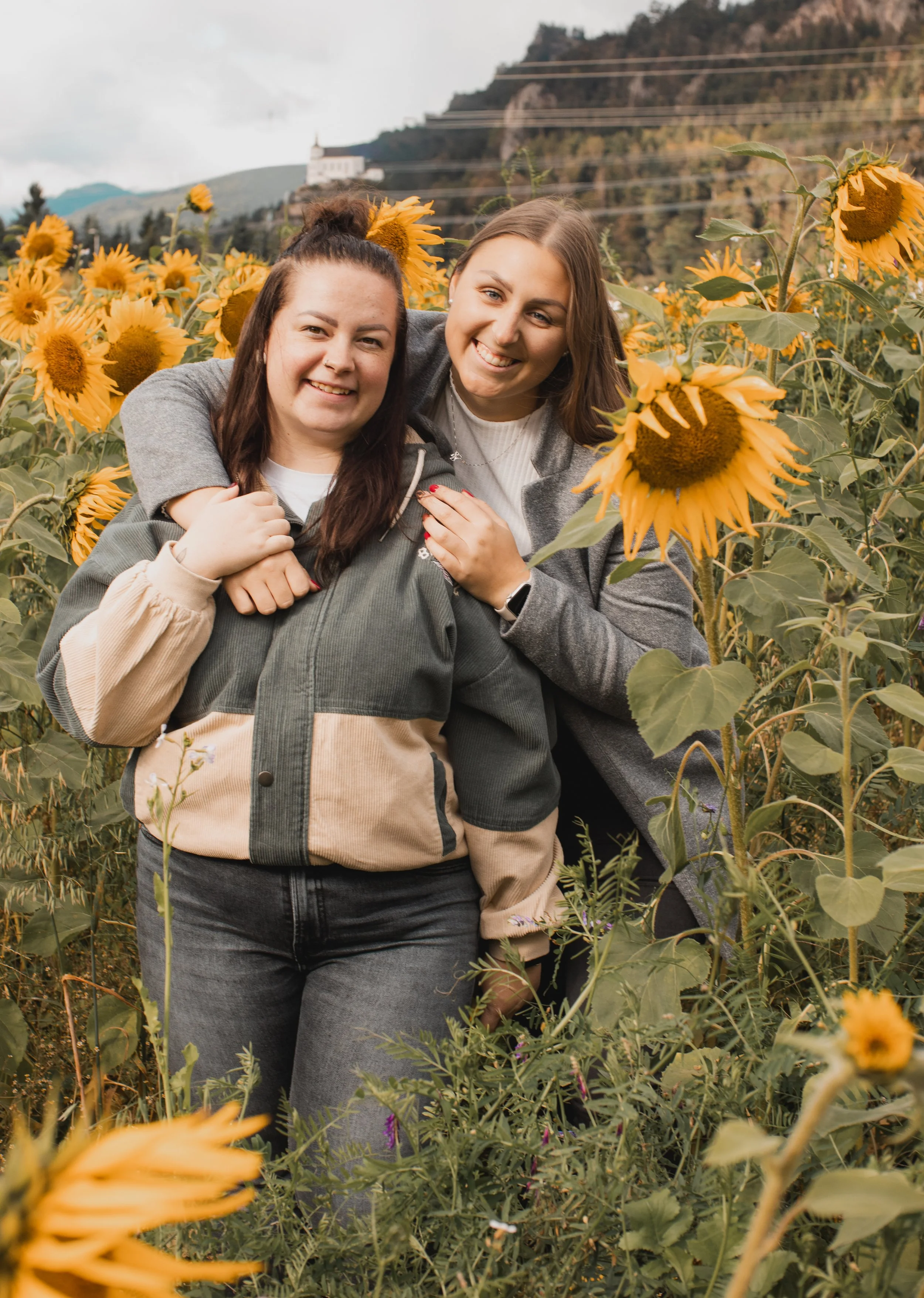 Zwei lachende Frauen in einem Sonnenblumenfeld, umgeben von hohen Sonnenblumen, mit Hügeln im Hintergrund.