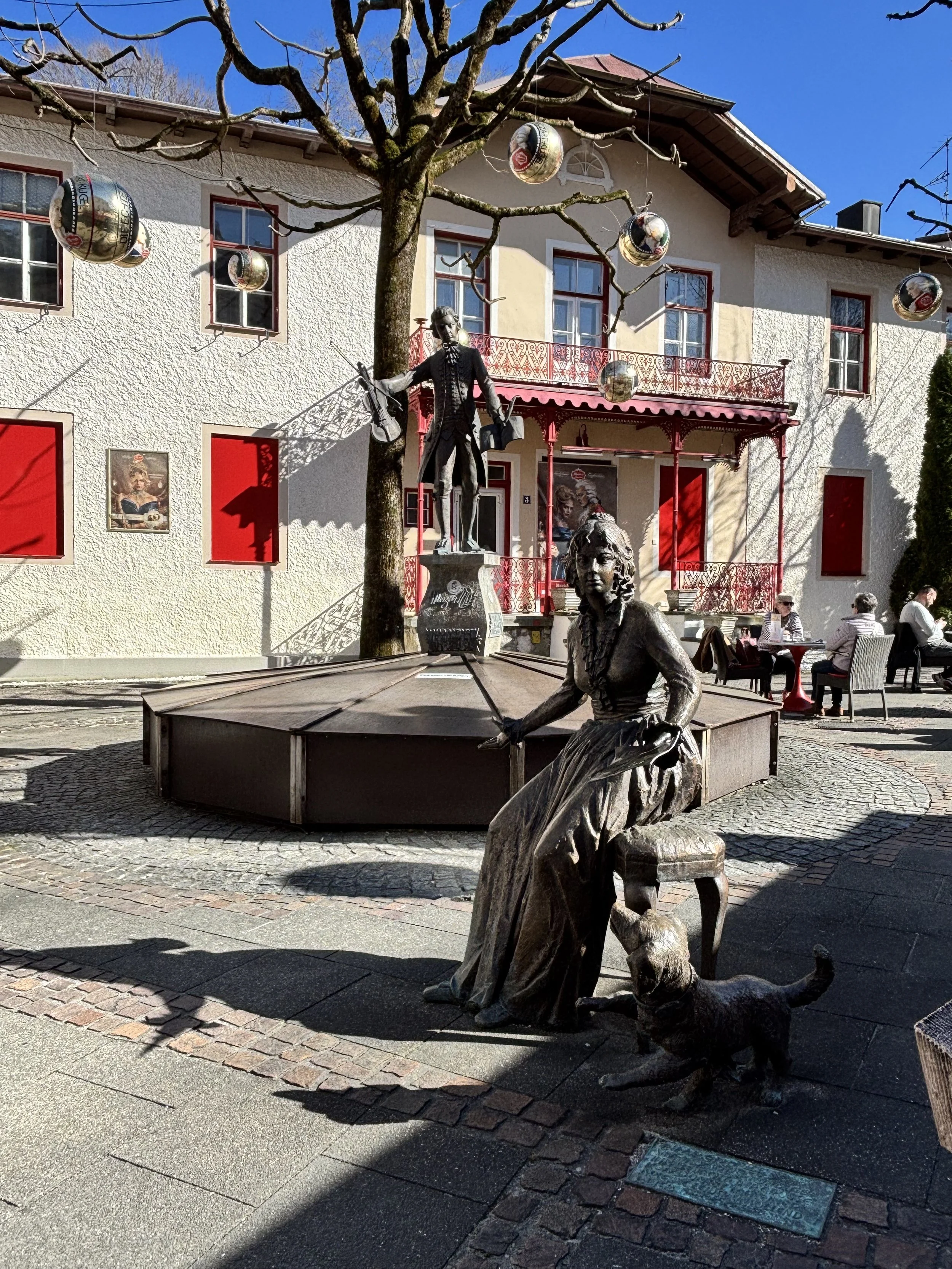 Denkmal mit drei Statuen vor einem weiß-blauen Gebäude mit roten Fensterläden und Balkon, umgeben von Bäumen und Sitzgelegenheiten, im Freien bei sonnigem Wetter.