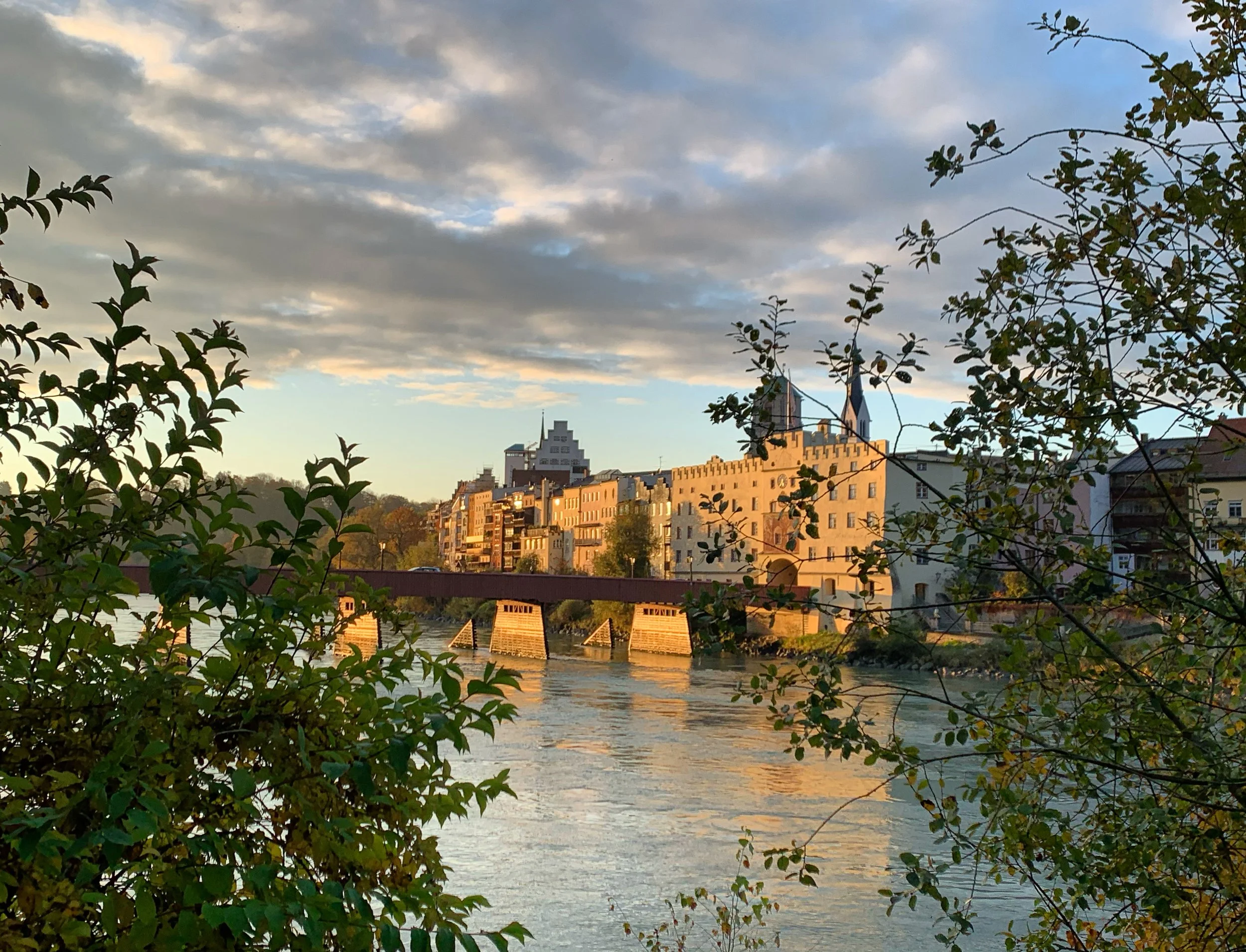 Wasserburg: Stadtansicht mit Fluss, Brücke und Gebäuden bei Sonnenuntergang, teilweise im Schatten durch Bäume im Vordergrund.