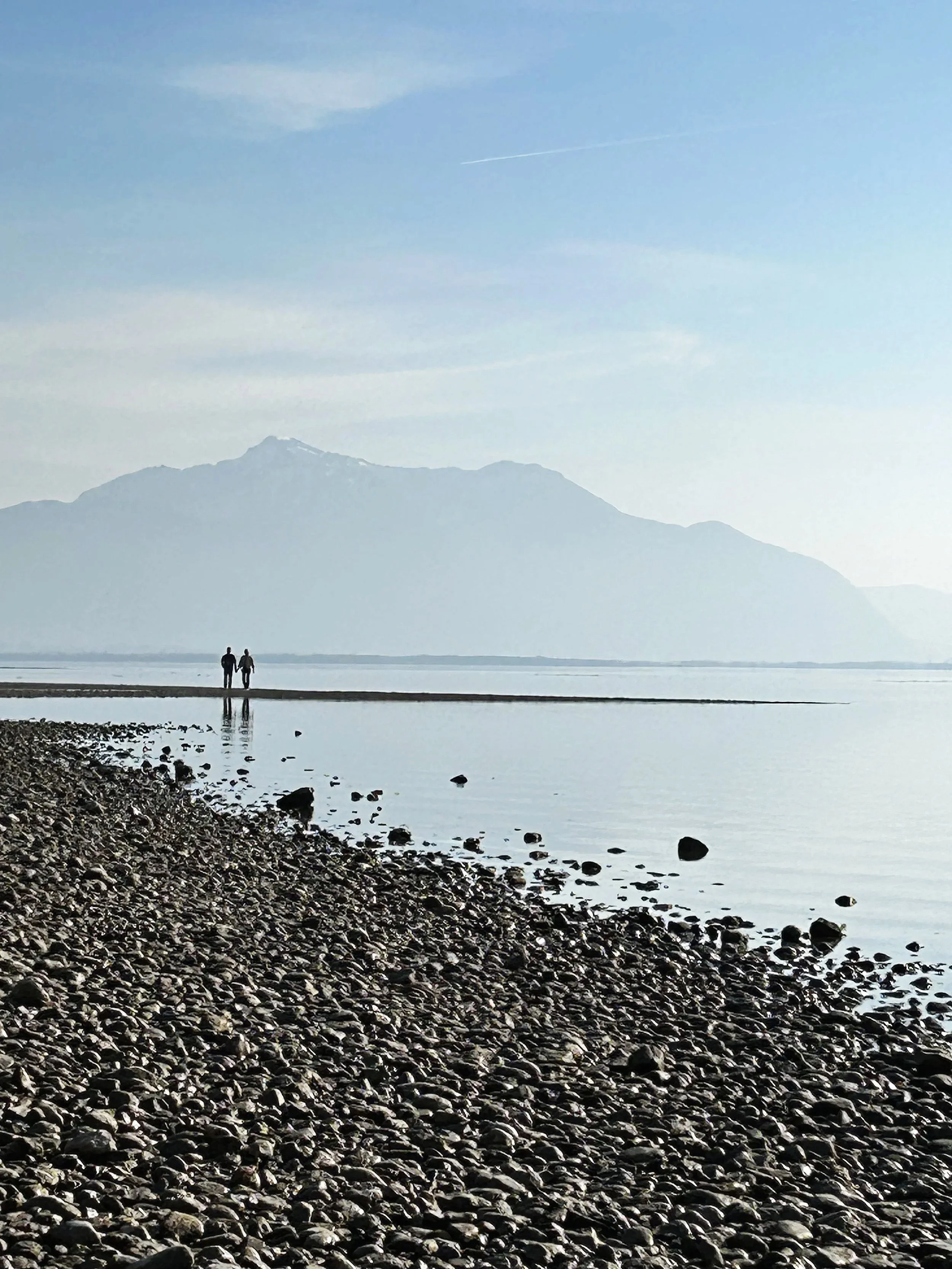 Zwei Personen stehen am Ufer des Chiemsees mit Bergen im Hintergrund, auf einem flachen Landstück. Der Himmel ist leicht bewölkt.
