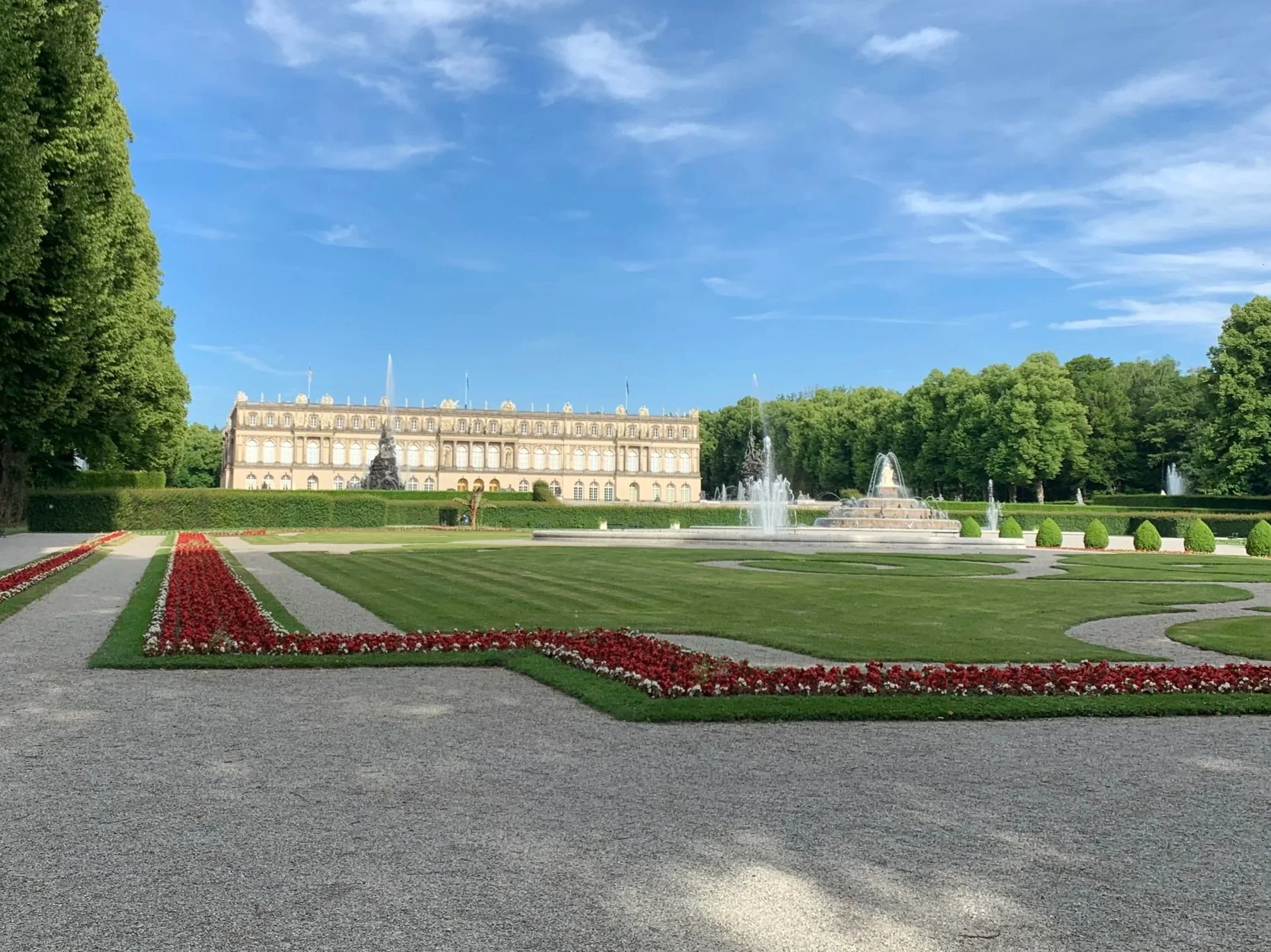 Ein großer Park mit gepflegtem Rasen, Blumenbeeten und Springbrunnen vor einem historischen Gebäude unter einem blauen Himmel.