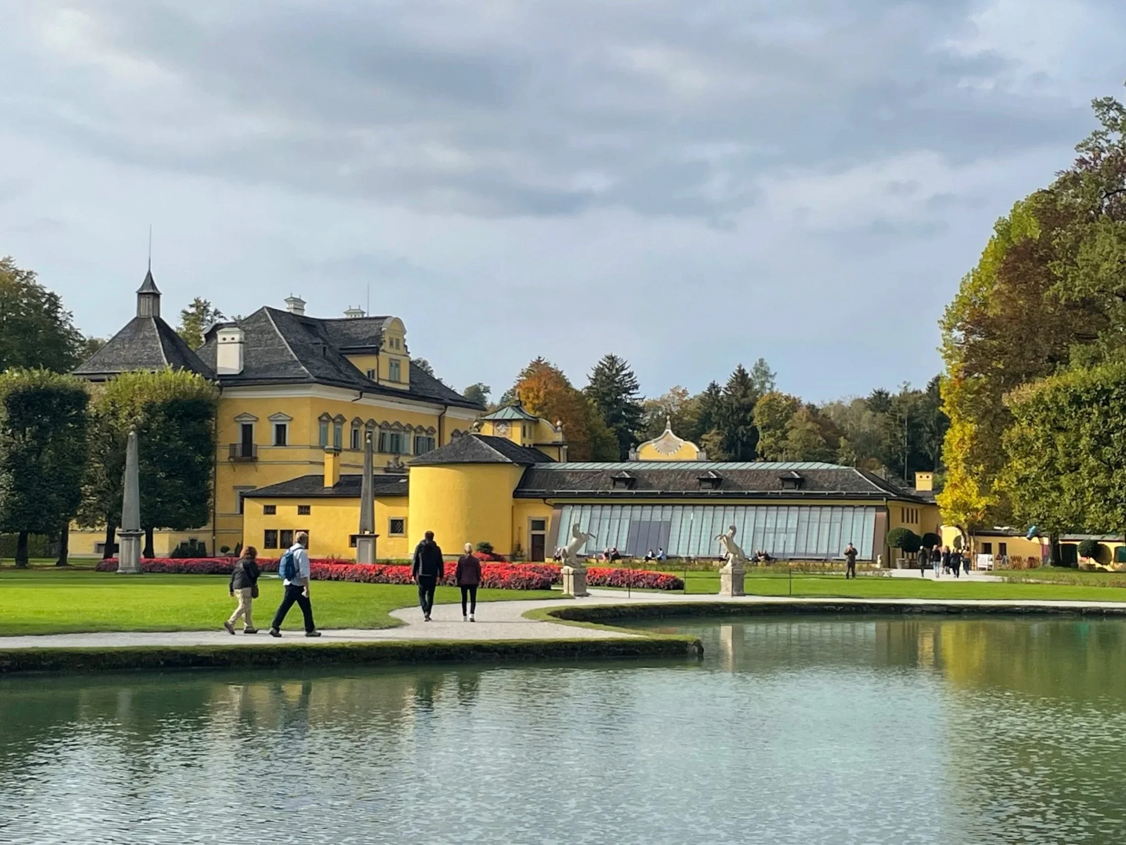 Historisches gelbes Gebäude mit Türmchen und großen Fenstern, umgeben von Bäumen, am Wasser mit Menschen beim Spaziergang
