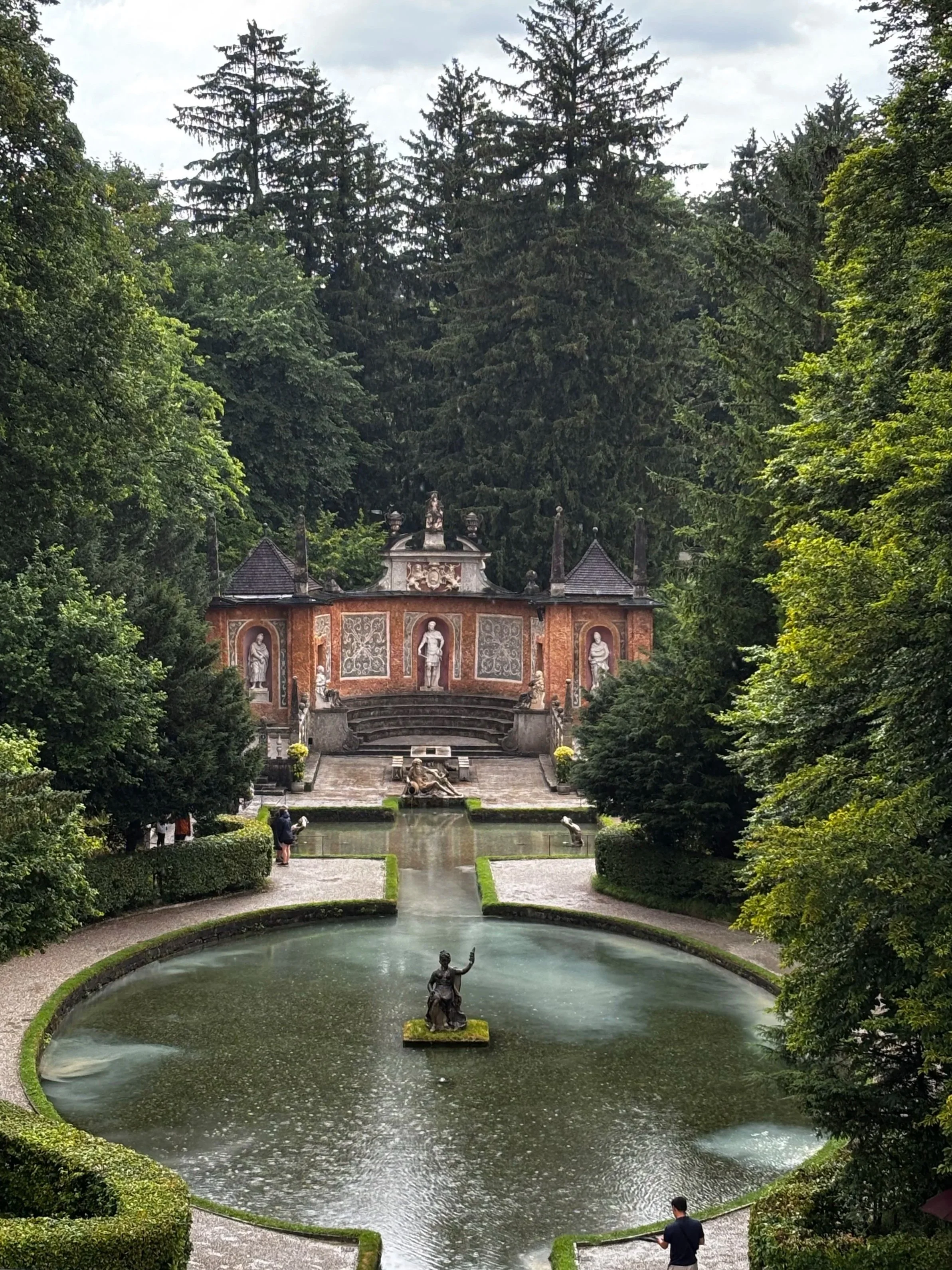 Ein Garten mit einem kreisförmigen Wasserbecken, in der Mitte eine Statue, im Hintergrund eine barockartige Struktur mit Skulpturen, umgeben von Bäumen.