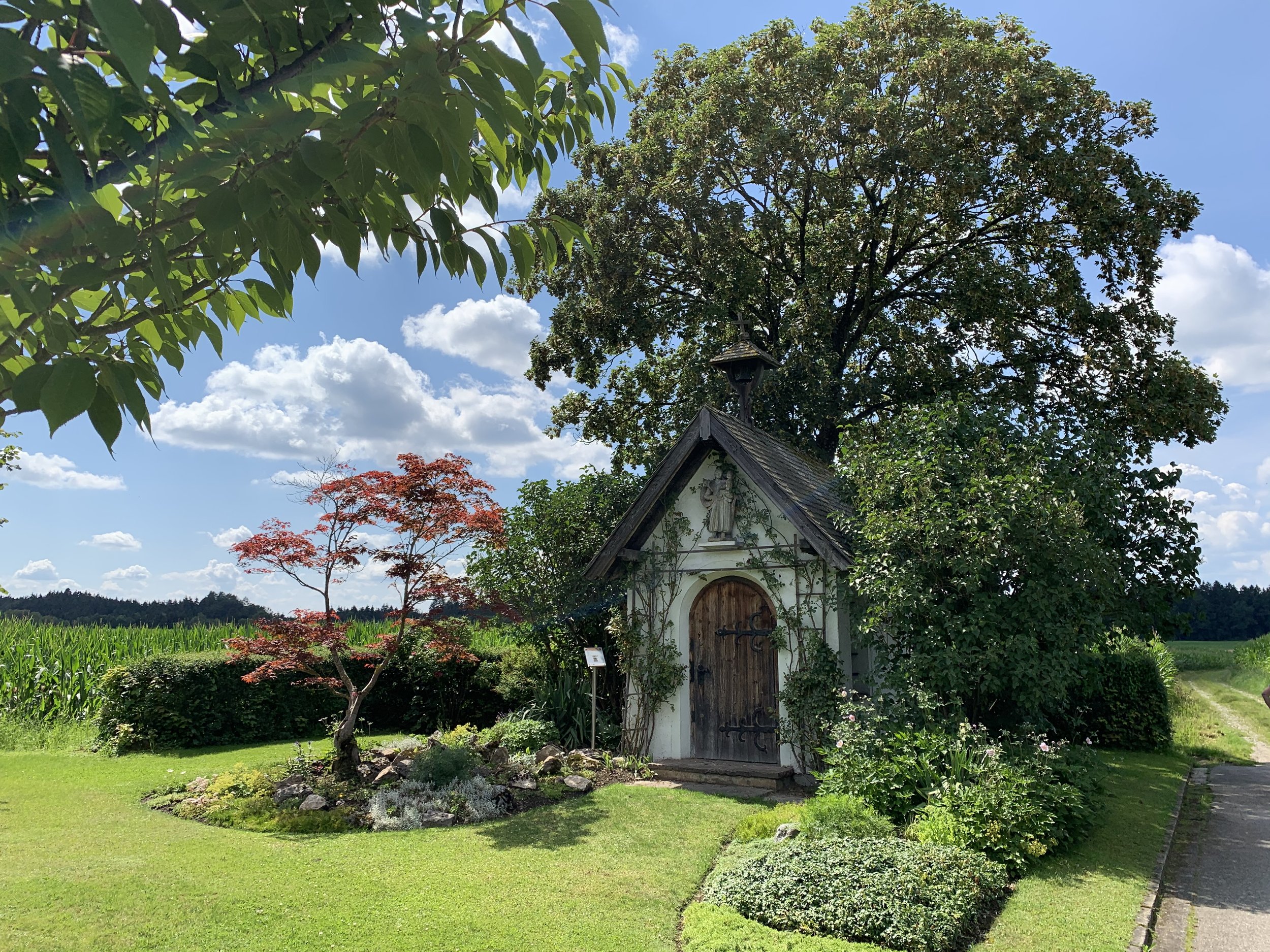 Ein kleiner Holzschuppen mit einer religiösen Skulptur an der Vorderseite, umgeben von grünen Büschen und Bäumen auf einer Wiese, mit einem klaren blauen Himmel.