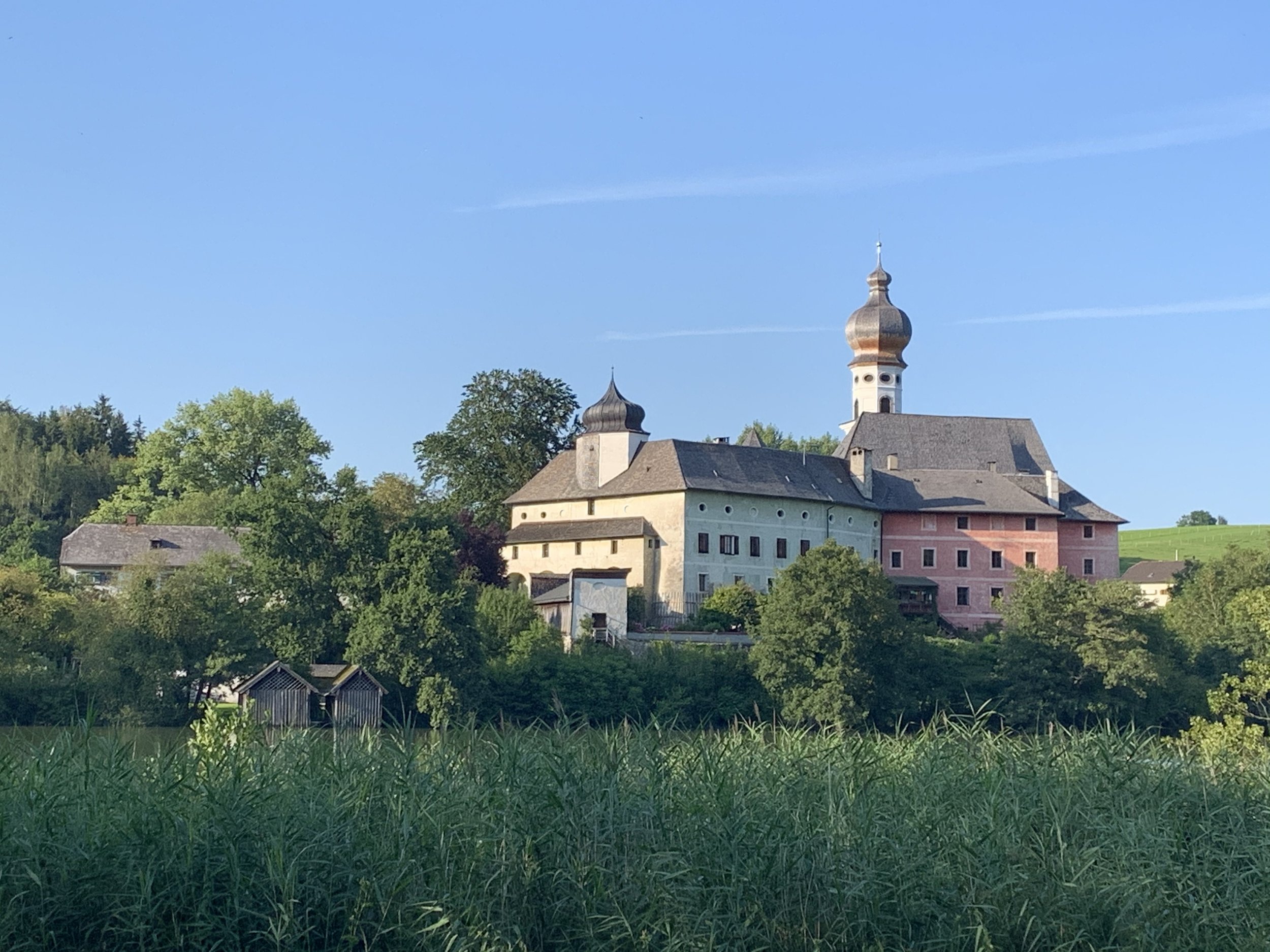 Blick auf eine historische Kirche auf einem Hügel, umgeben von Bäumen und Wiesen, bei sonnigem Himmel.