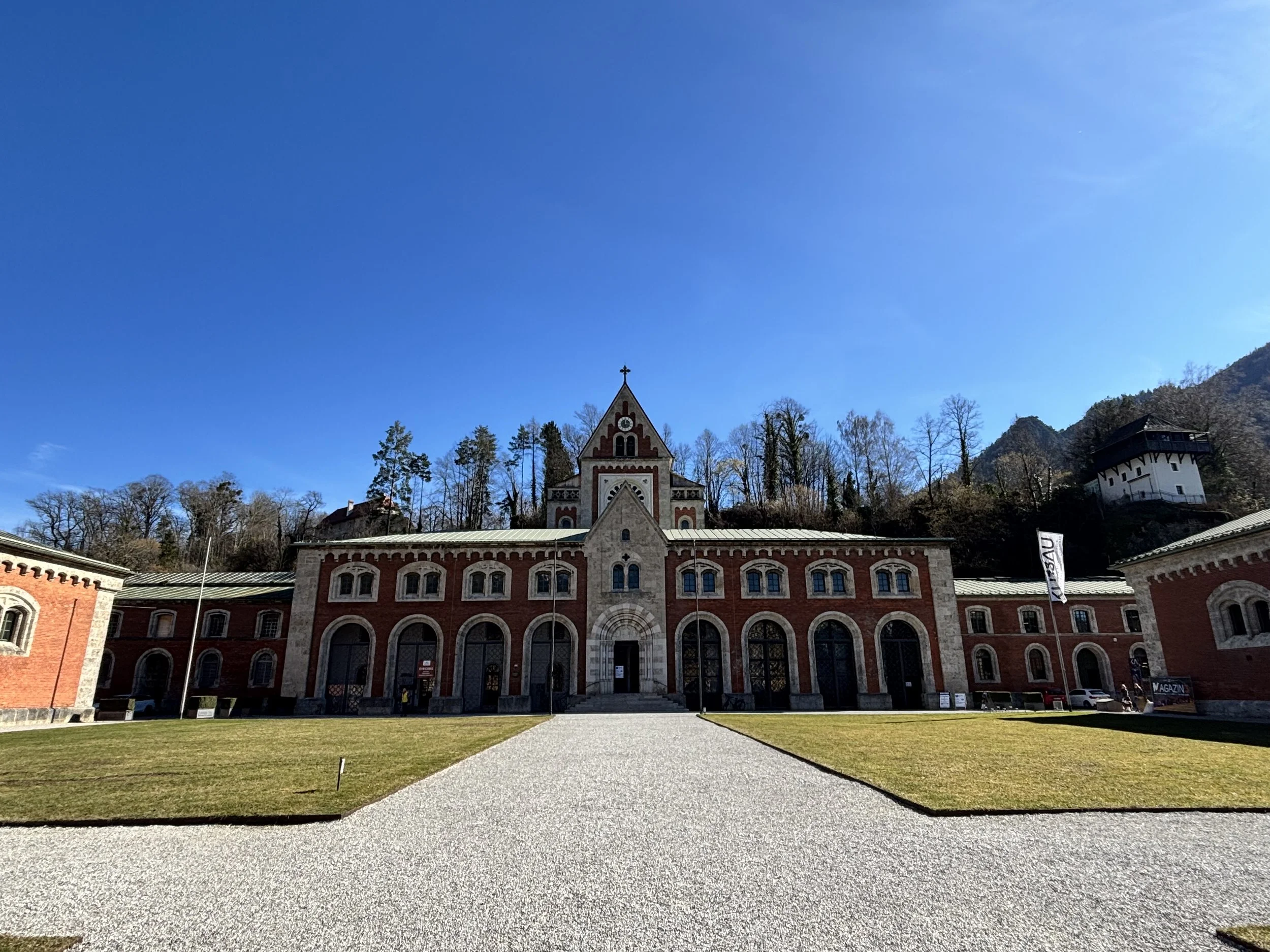 Historisches Gebäude mit rotem Backstein und arched Fenstern vor einem blauen Himmel, umgeben von Bäumen auf einem Hügel.