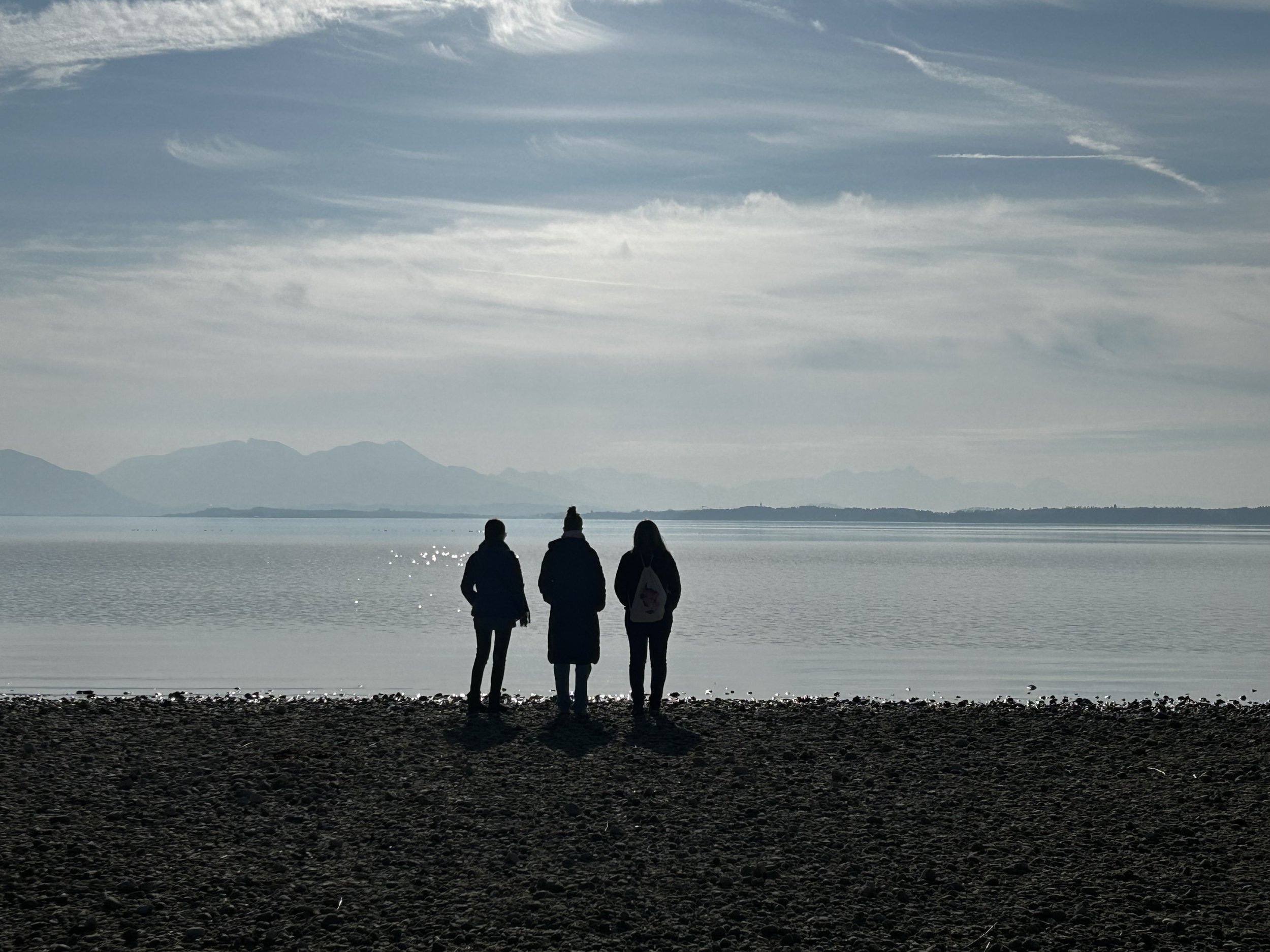 Drei Personen stehen am Ufer eines Sees und schauen auf das Wasser, im Hintergrund sind Berge und Wolken am Himmel sichtbar.