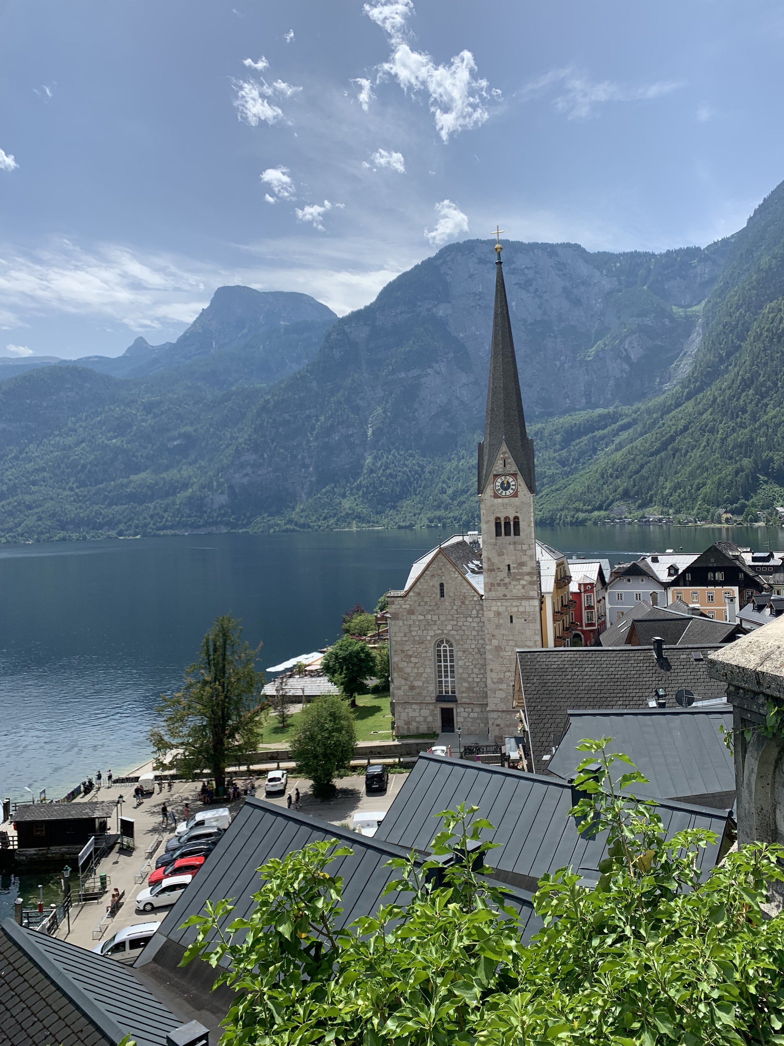 Blick auf ein historisches Gebäude mit Kirchturm an einem See inmitten von grünen Bergen, mit Parkplätzen und Bäumen im Vordergrund, bei schönem Wetter.