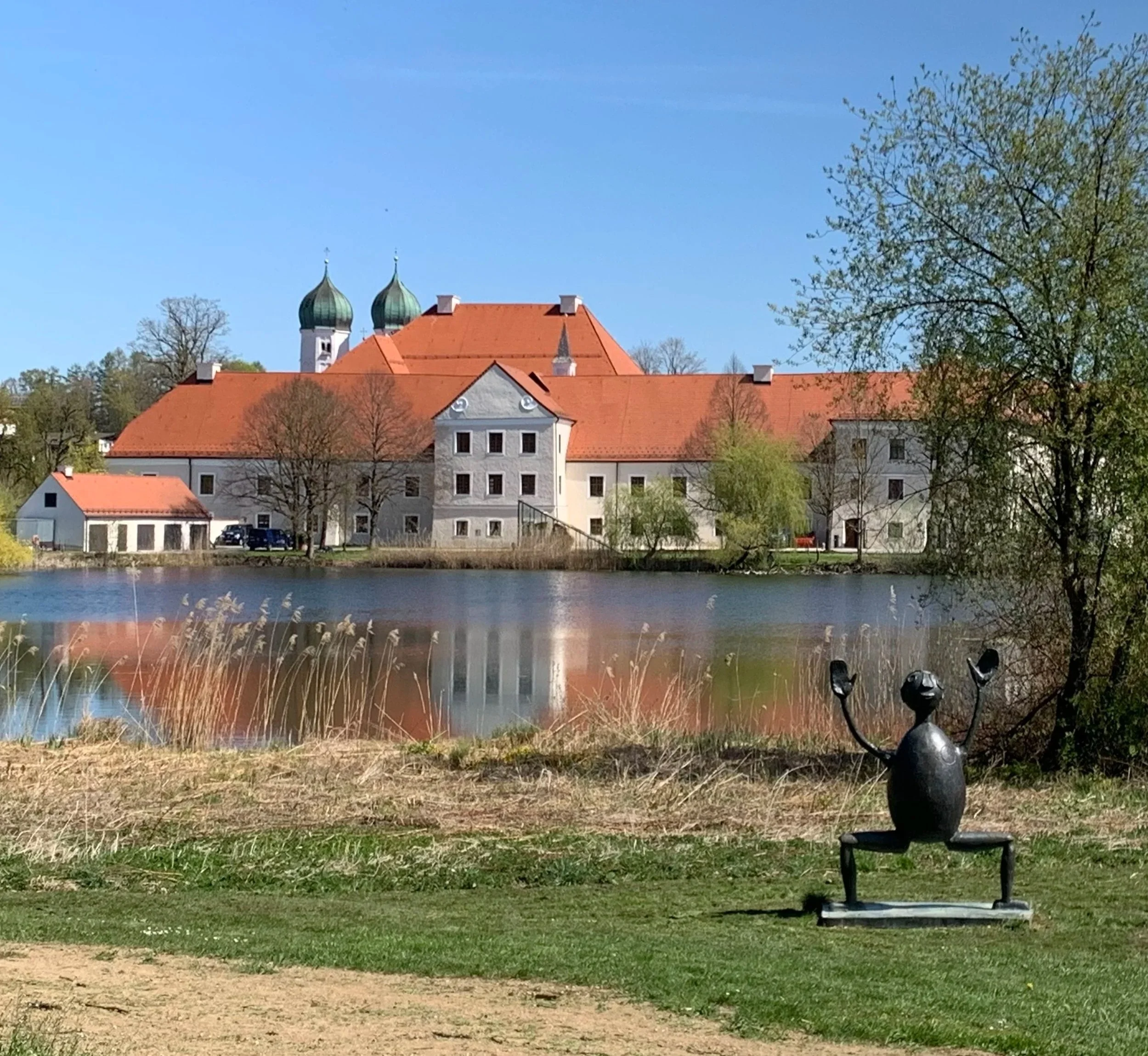 Blick auf Kloster Seeon, im Vordergrund eine Bronzeskulptur, umgeben von Gräsern und Bäumen, bei sonnigem Wetter.