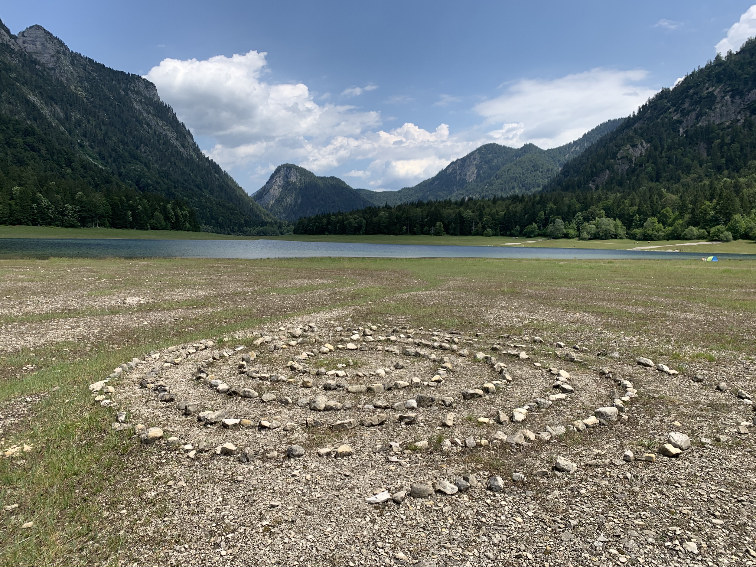 Ein Kreis aus Steinen auf trockenem Boden vor einem See mit Bergen im Hintergrund, bei blauem Himmel mit Wolken.