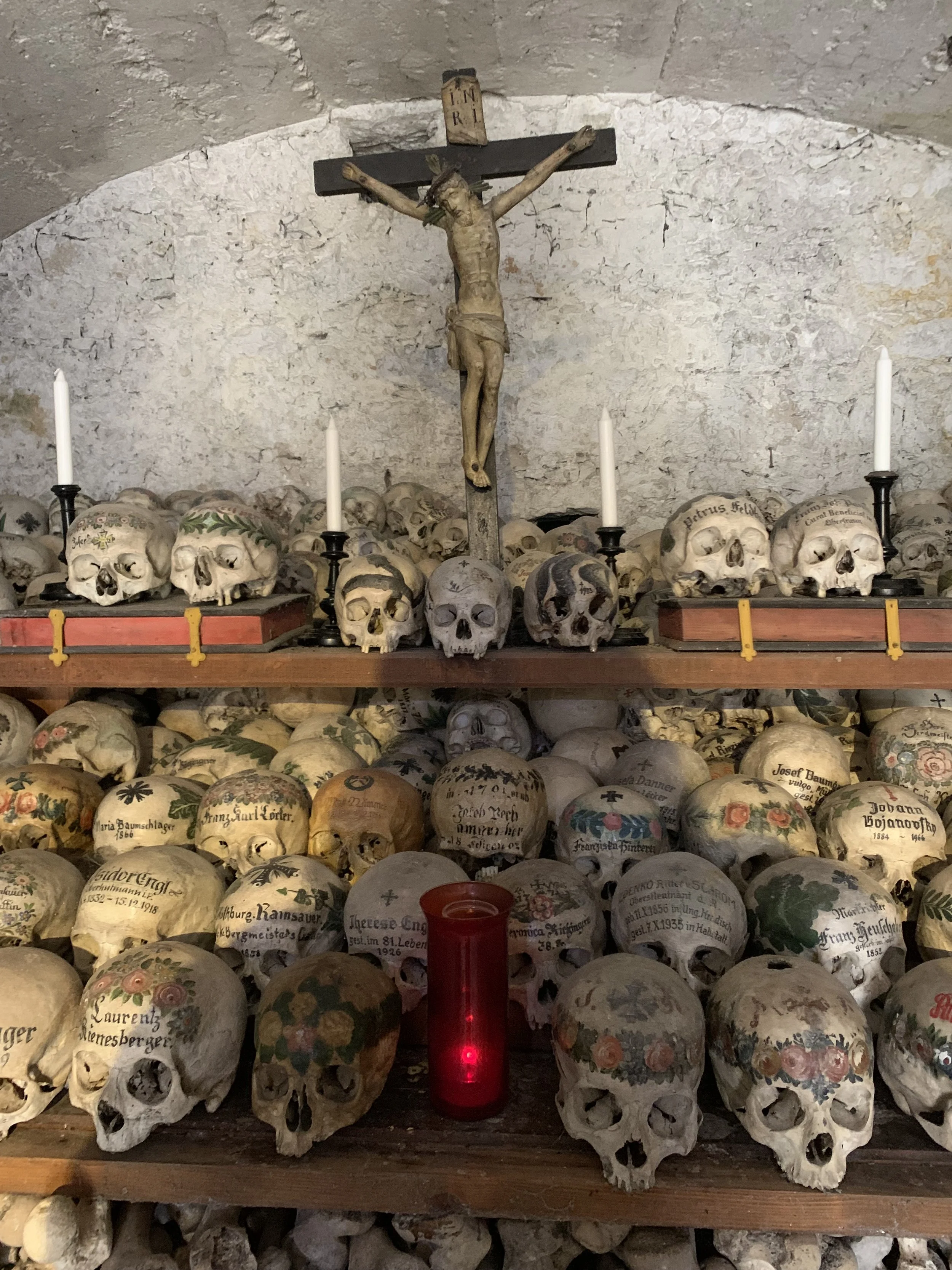 Ein Altar mit zahlreichen menschlichen Totenköpfen, einer Holz-Kruzifix-Statue Jesus Christus, vier weiße Kerzen und einen roten Kerzenhalter in einer alten, steinernen Umgebung.