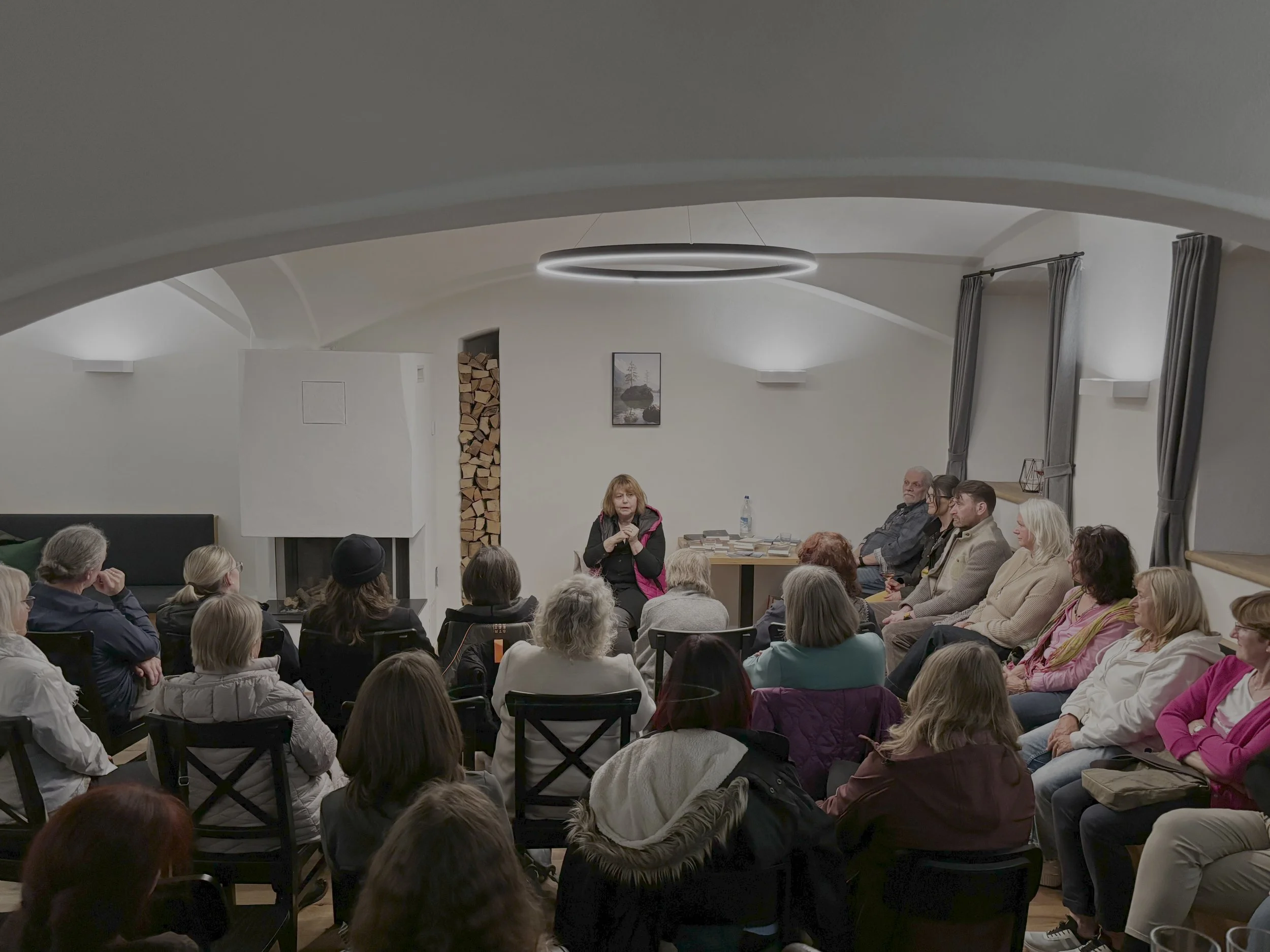Lona Pircha-Höberth spricht vor einer Gruppe von Menschen in einem hellen Raum mit Kamin und Holzstapel im Hintergrund.