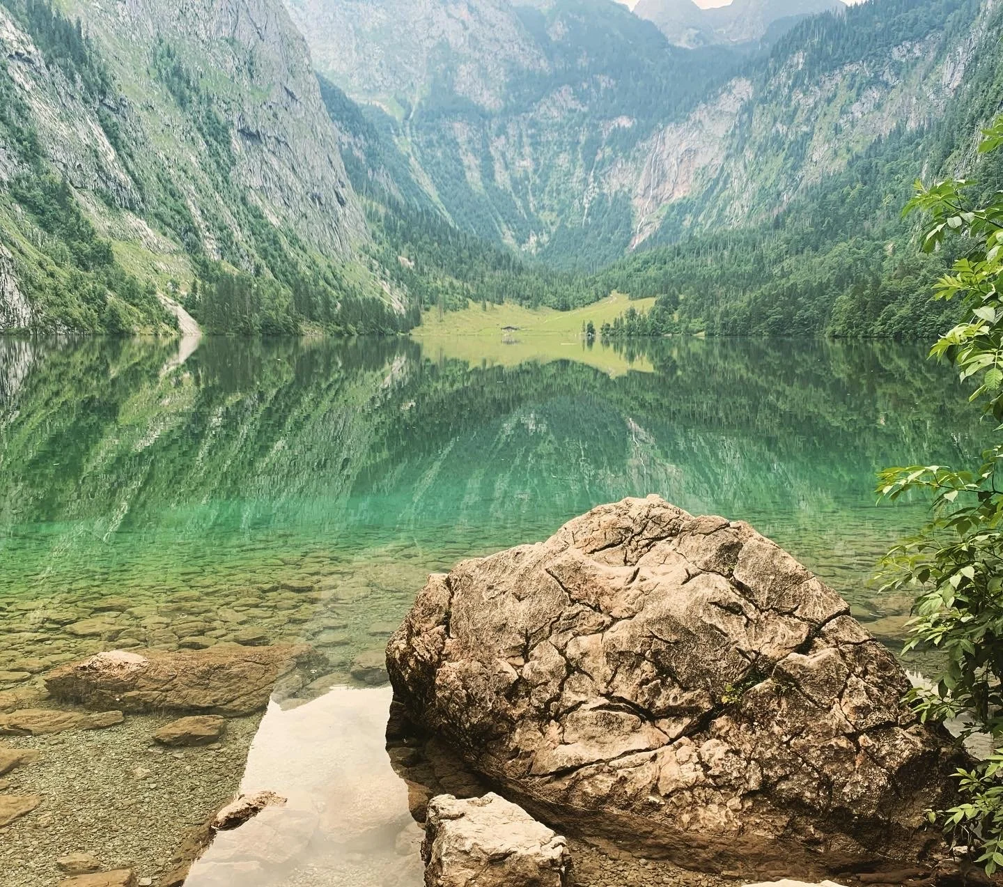 Ein ruhiger See inmitten von Bergen mit klarem Wasser, reflektiert die umliegende grüne Landschaft und Felsen im Vordergrund.