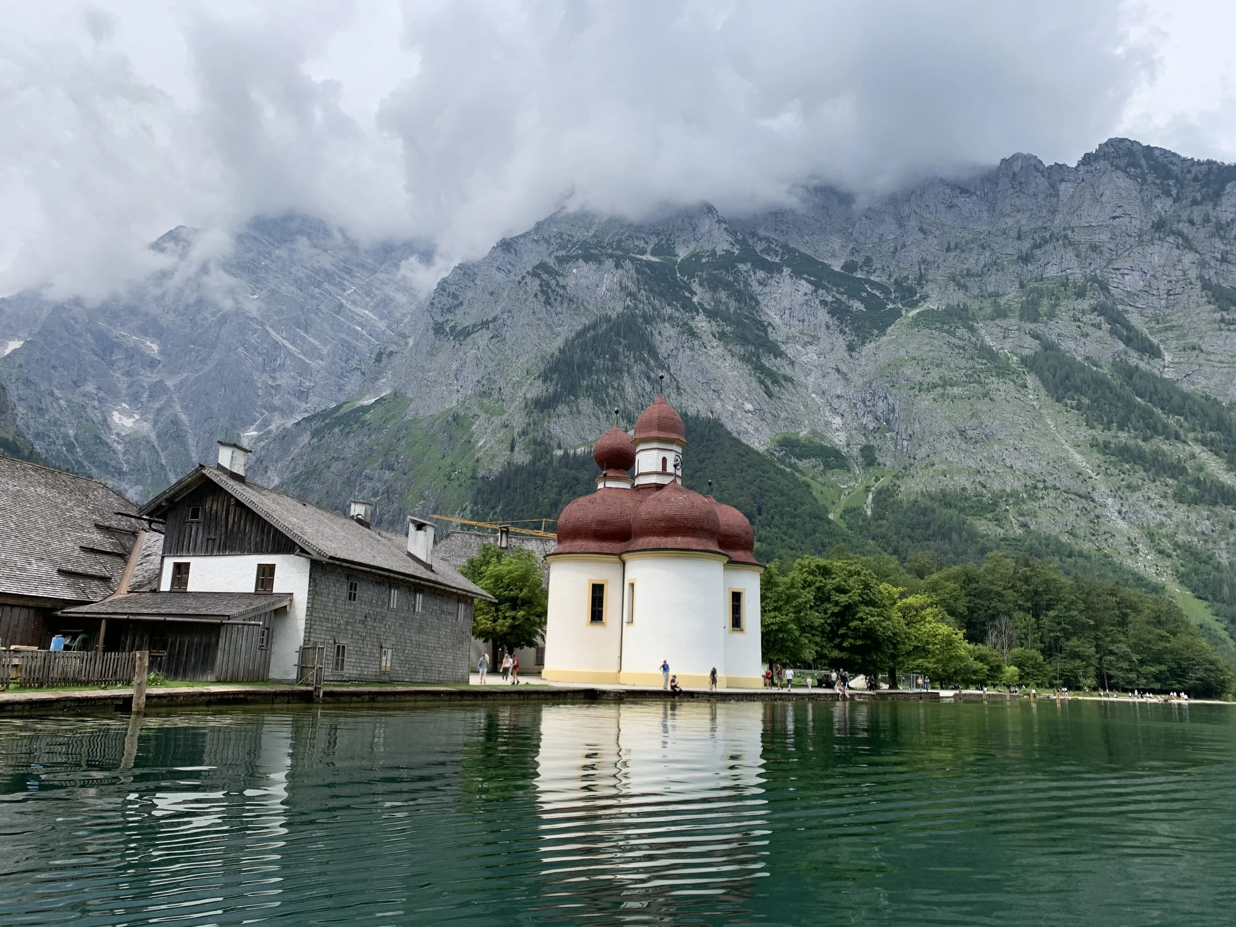 Ein kleines weißes Kirche mit roten Kuppeln am Ufer eines Sees, umgeben von Bäumen und Bergen im Hintergrund, teilweise von Wolken bedeckt.