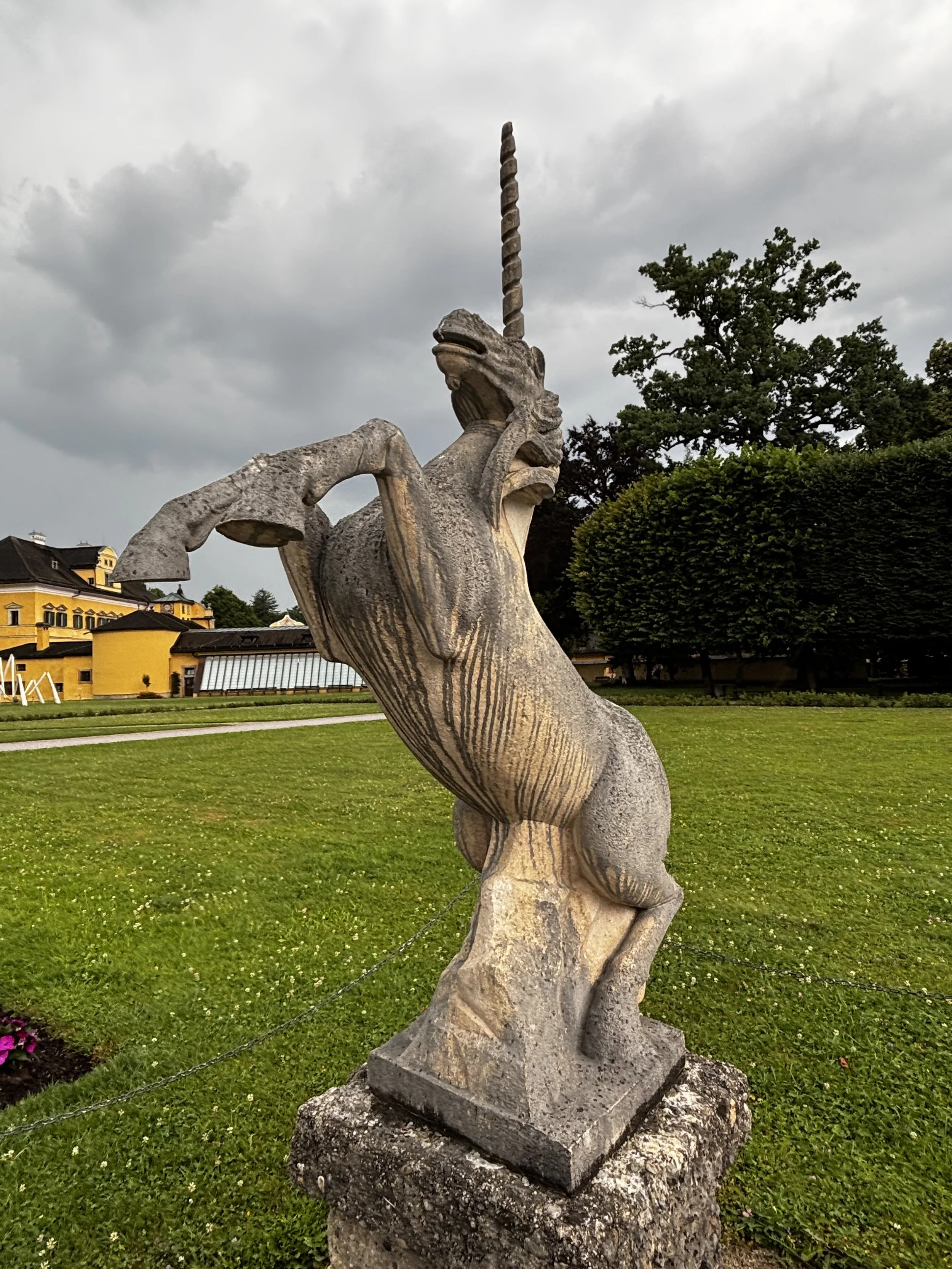 Skulptur eines Einhorns in einem Park mit grüner Wiese und Bäumen im Hintergrund bei wolkigem Himmel.