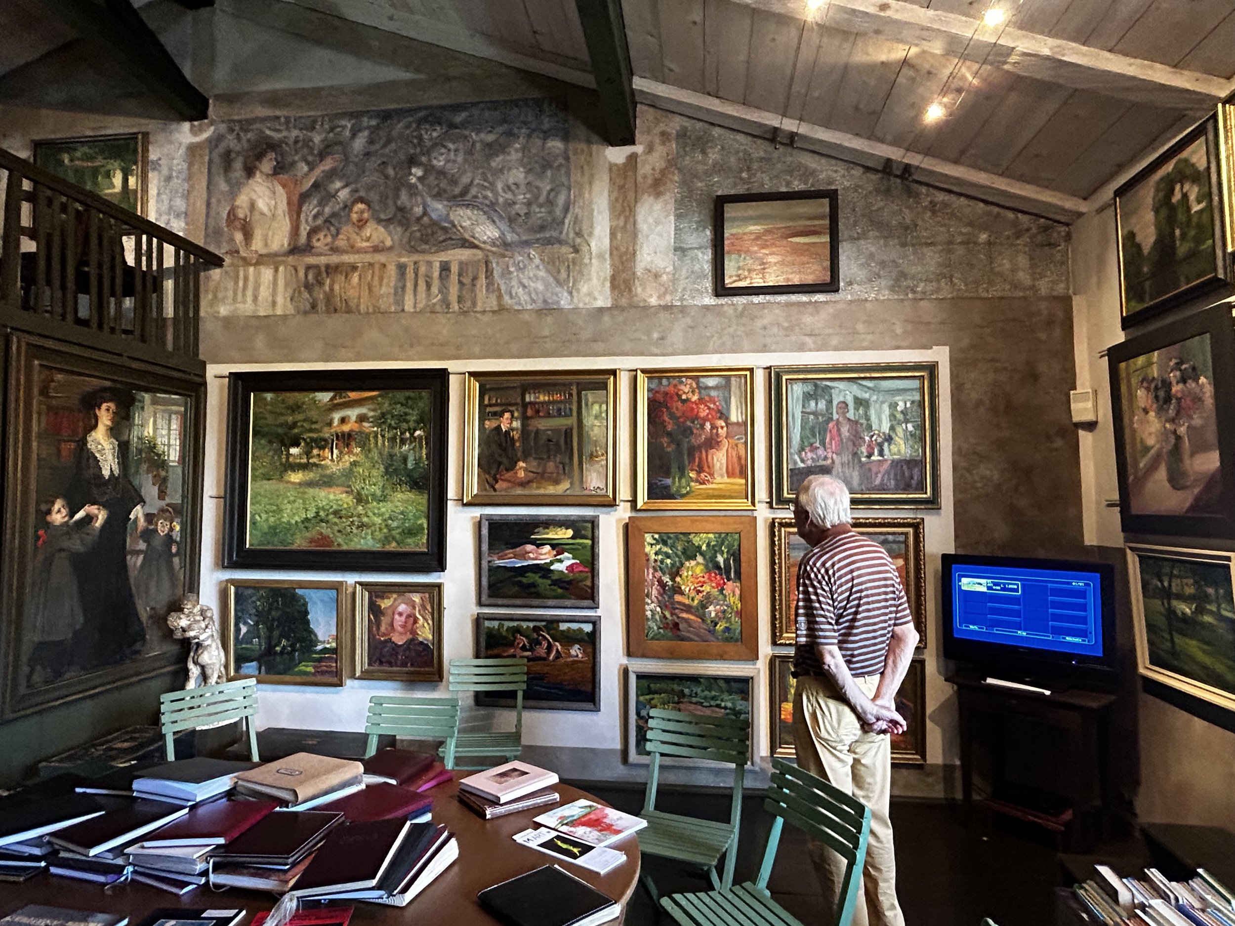 Ein älterer Mann betrachtet Kunstwerke in einer Galerie, die an einer Wand hängen, umgeben von weiteren Gemälden und Skulpturen, mit einem Tisch voller Bücher im Vordergrund.