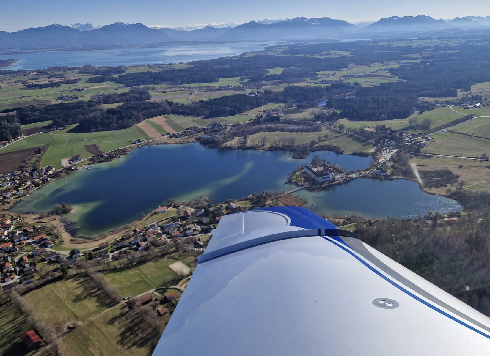 Luftaufnahme von einem Flugzeug in der Luft, das über eine Landschaft mit Seen, Feldern und Bergen fliegt.