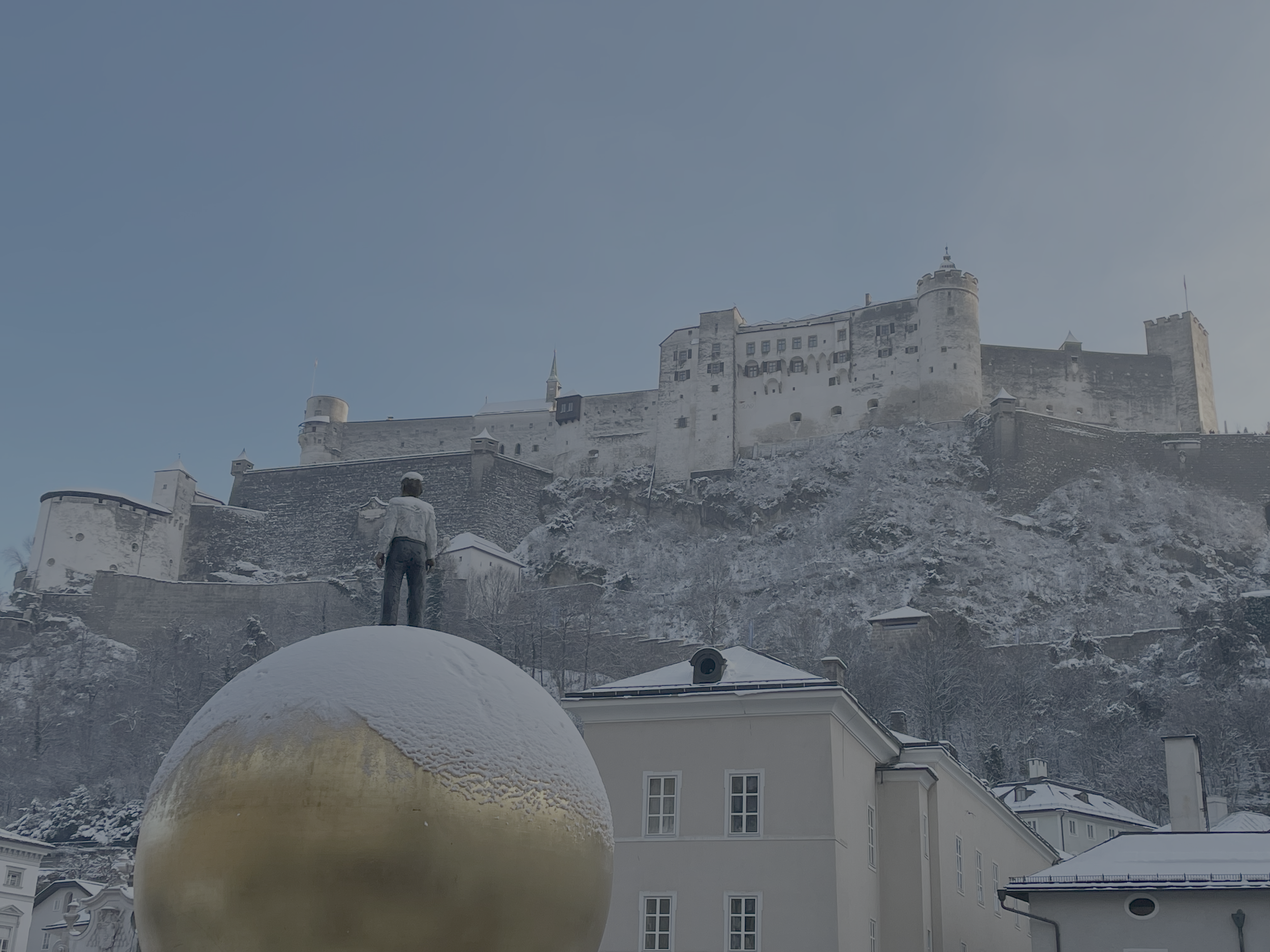 Burg auf einem Hügel im Winter, mit Schneefall, ein Mensch steht vor einer großen goldenen Kugel im Vordergrund.