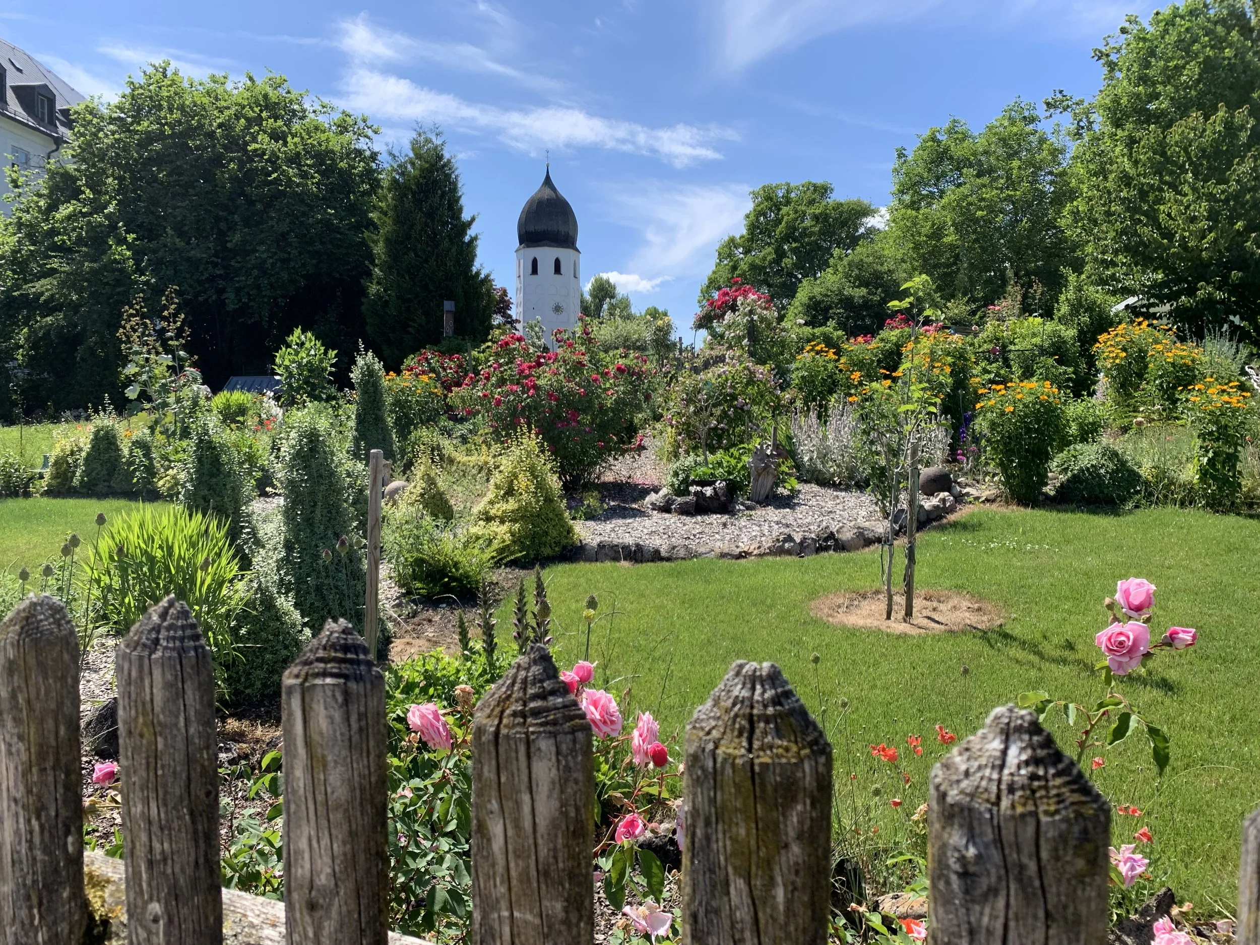 Fraueninsel: Blick auf einen gepflegten Garten mit Blumenbeeten, einer kleinen Baumgruppe, einer grünen Wiese und einer weißen Kirche mit dunklem Dach im Hintergrund, eingebettet in Landschaft mit Bäumen und blauen Himmel.