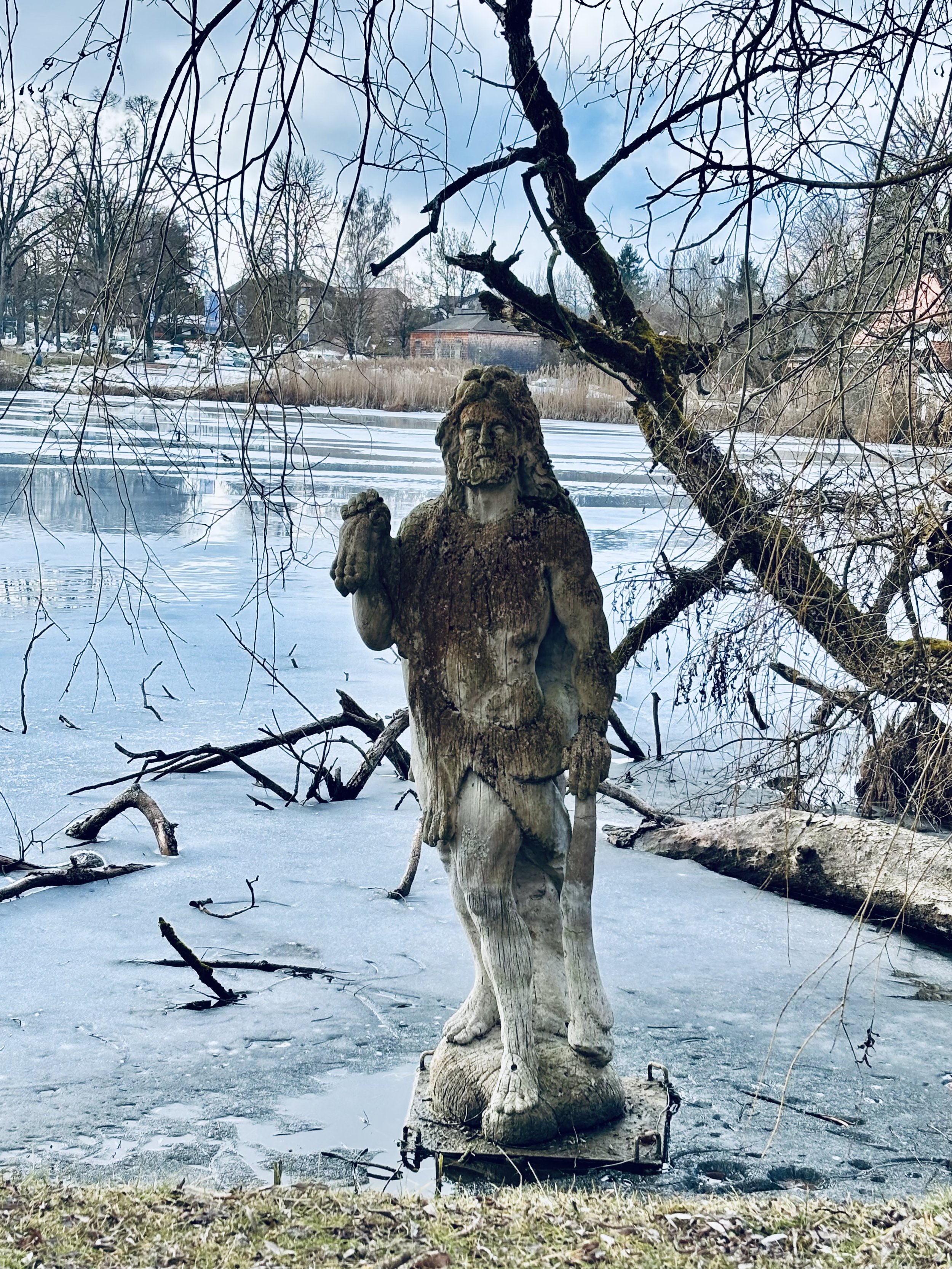 Eine Skulptur eines Löwen, die auf einem Sockel im Winter auf einer vereisten Wasserfläche steht, mit Bäumen und einem Ort im Hintergrund.