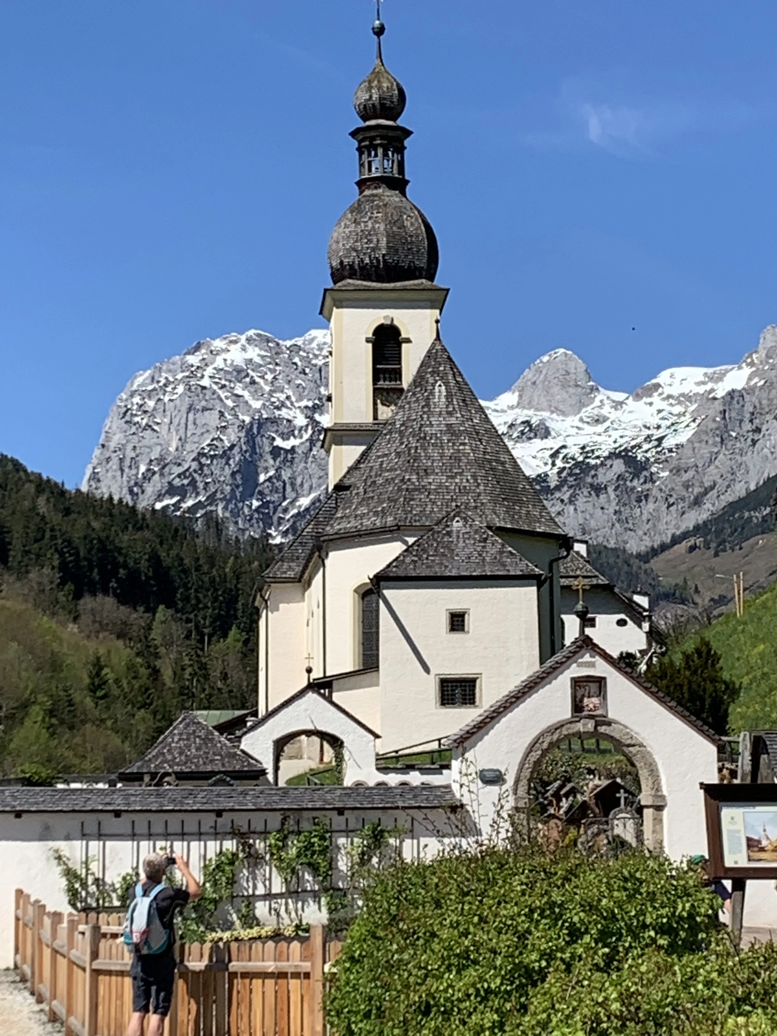 Ein kleines weißes Kirchengebäude mit dunklen Dächern vor schneebedeckten Bergen unter blauem Himmel, mit ein paar Menschen im Vordergrund.