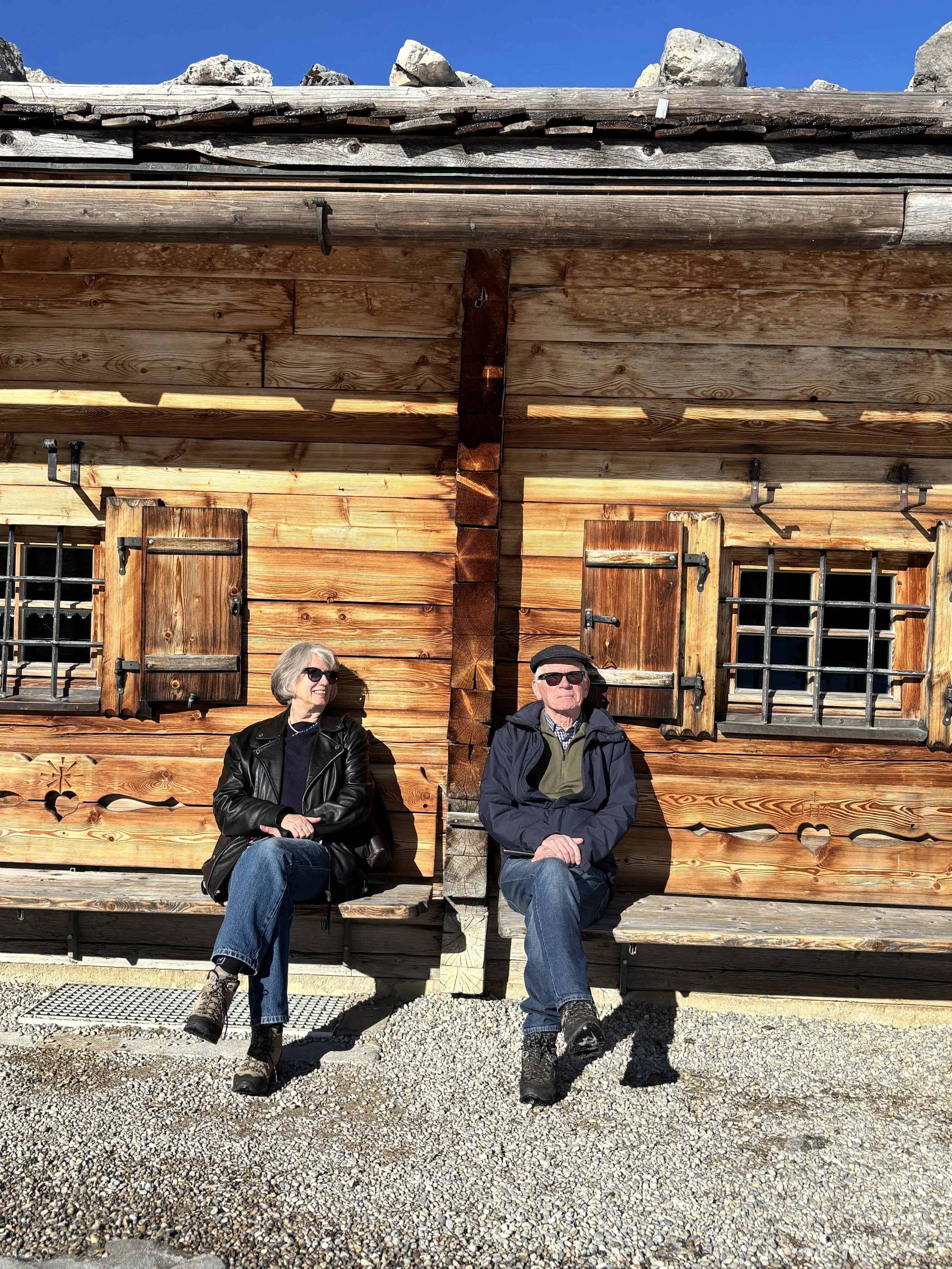 Ein älteres Paar sitzt auf einer Holzbank vor einer rustikalen Holzhütte mit kleinen Fensterläden und Gitterfenstern. Es ist sonnig, und sie tragen Sonnenbrillen und warme Kleidung.