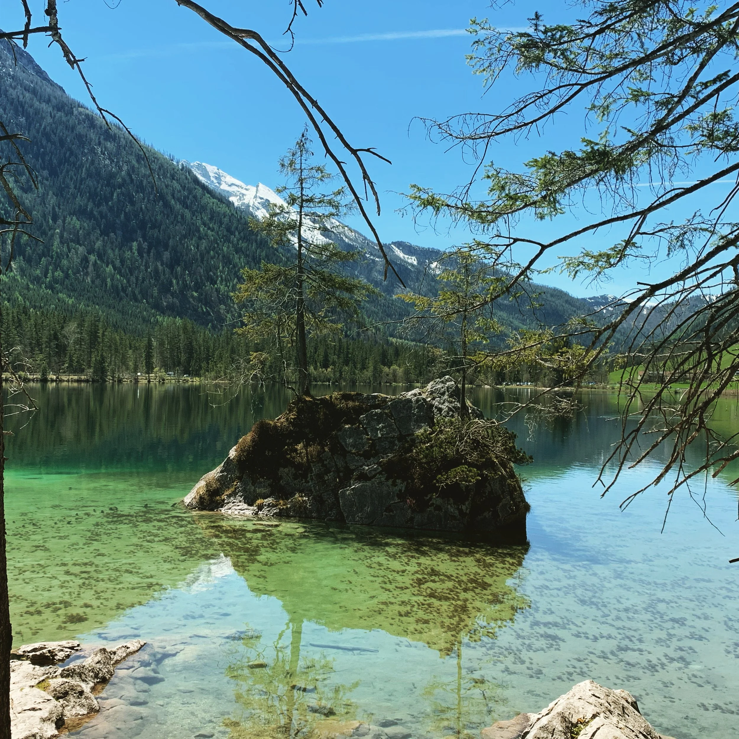 Ein klarer Bergsee mit einem großen Stein in der Mitte, umgeben von Bergen und Wäldern, mit blauer Himmel im Hintergrund.