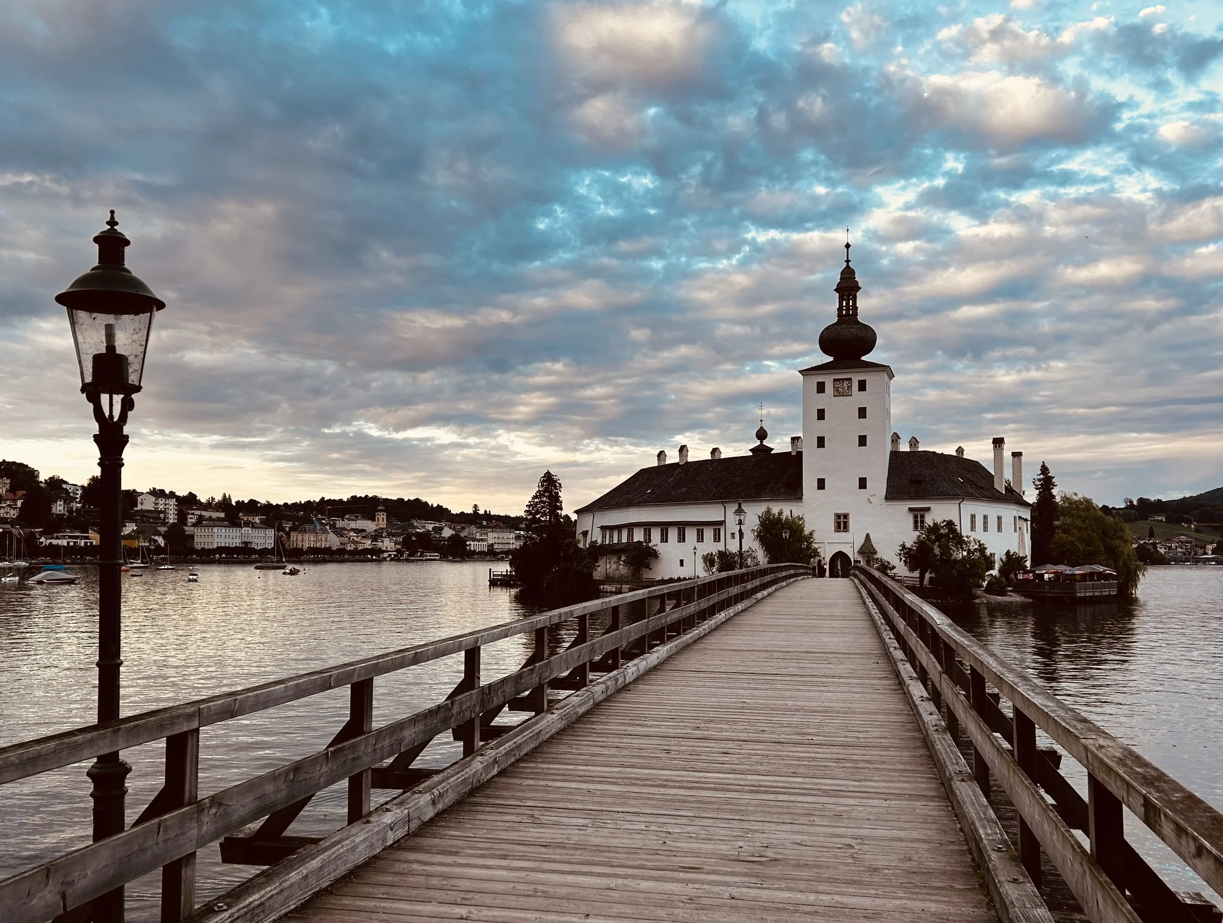 Ein Holzsteg führt zu einem weißen Schloss auf einer kleinen Insel im See, im Hintergrund sind Häuser und Bäume, der Himmel ist bewölkt bei Sonnenuntergang.