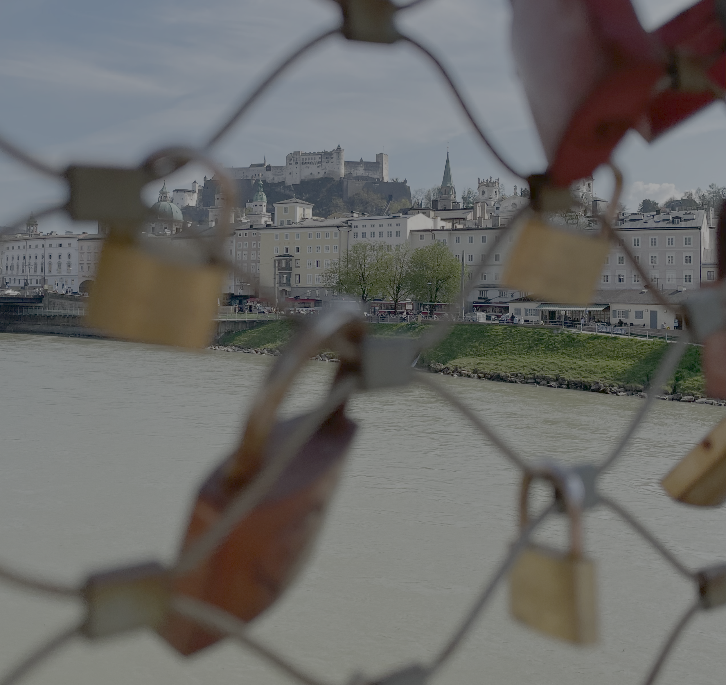 Blick auf die Stadt Salzburg mit Festung Hohensalzburg auf einem Hügel, sichtbar durch ein absichtlich unscharfes Gitter mit Liebesschlössern im Vordergrund, am Ufer des Flusses Salzach.