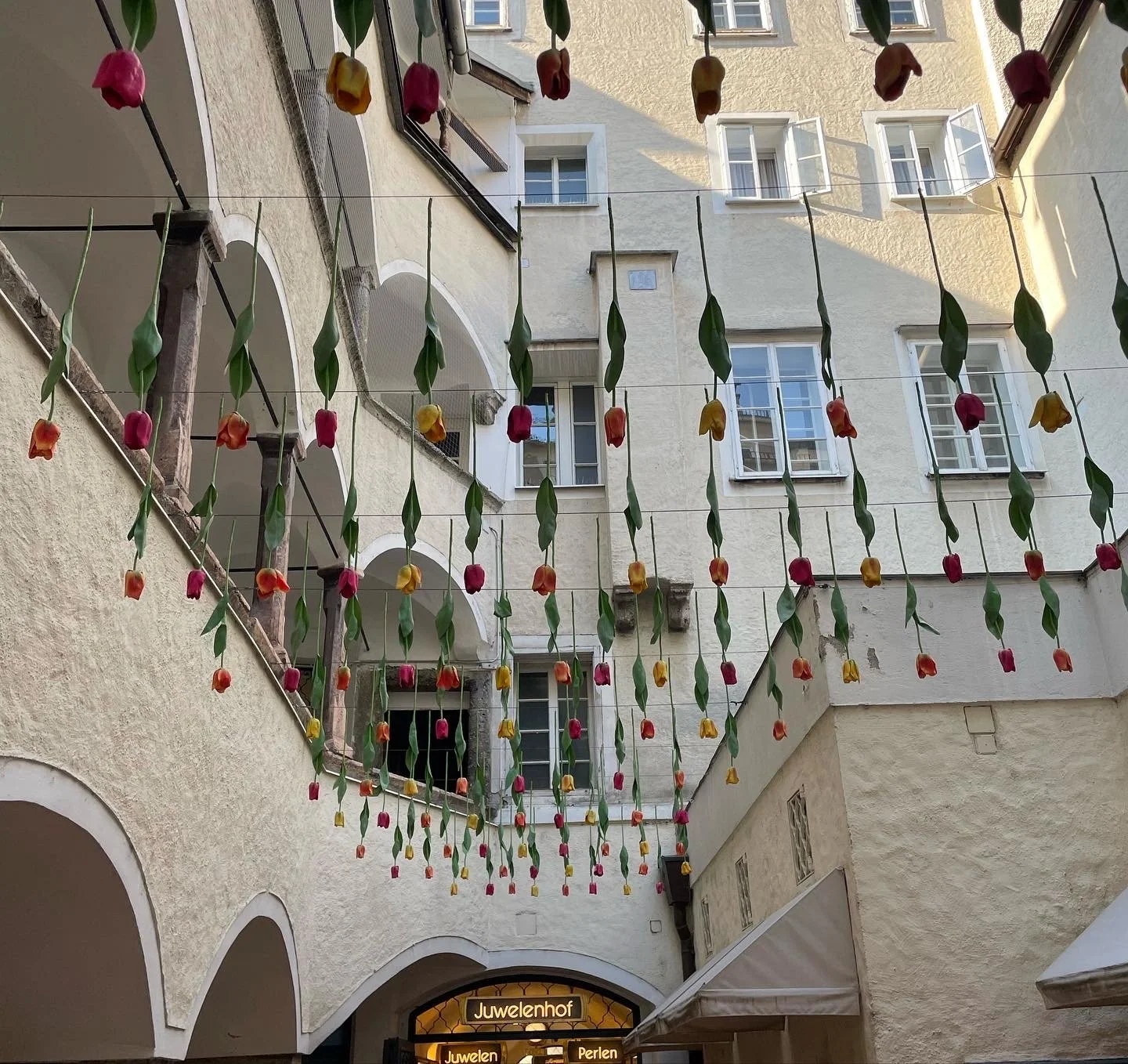 Blick auf einen Innenhof mit hängenden Dekorationen aus bunten Tulpen aus Papier oder Stoff, einem Geschäftsschild 'Juwelenhof' und offenen Fenstern an den umliegenden Gebäuden.