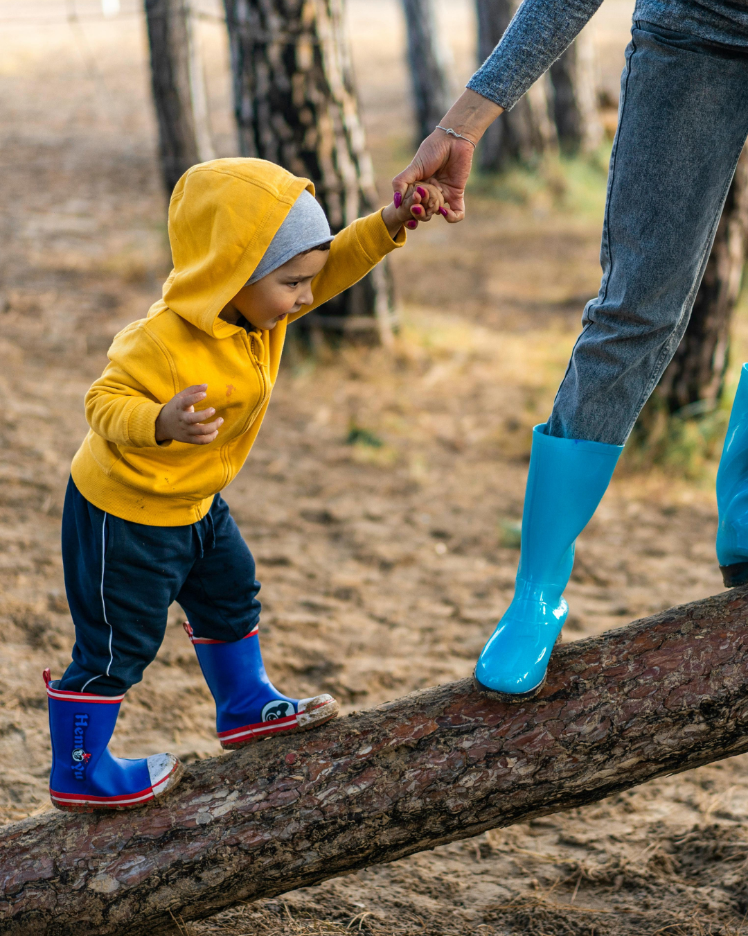 A young child wearing a yellow hoody and blue wellies climbing up a log while holding on to an adults hand.