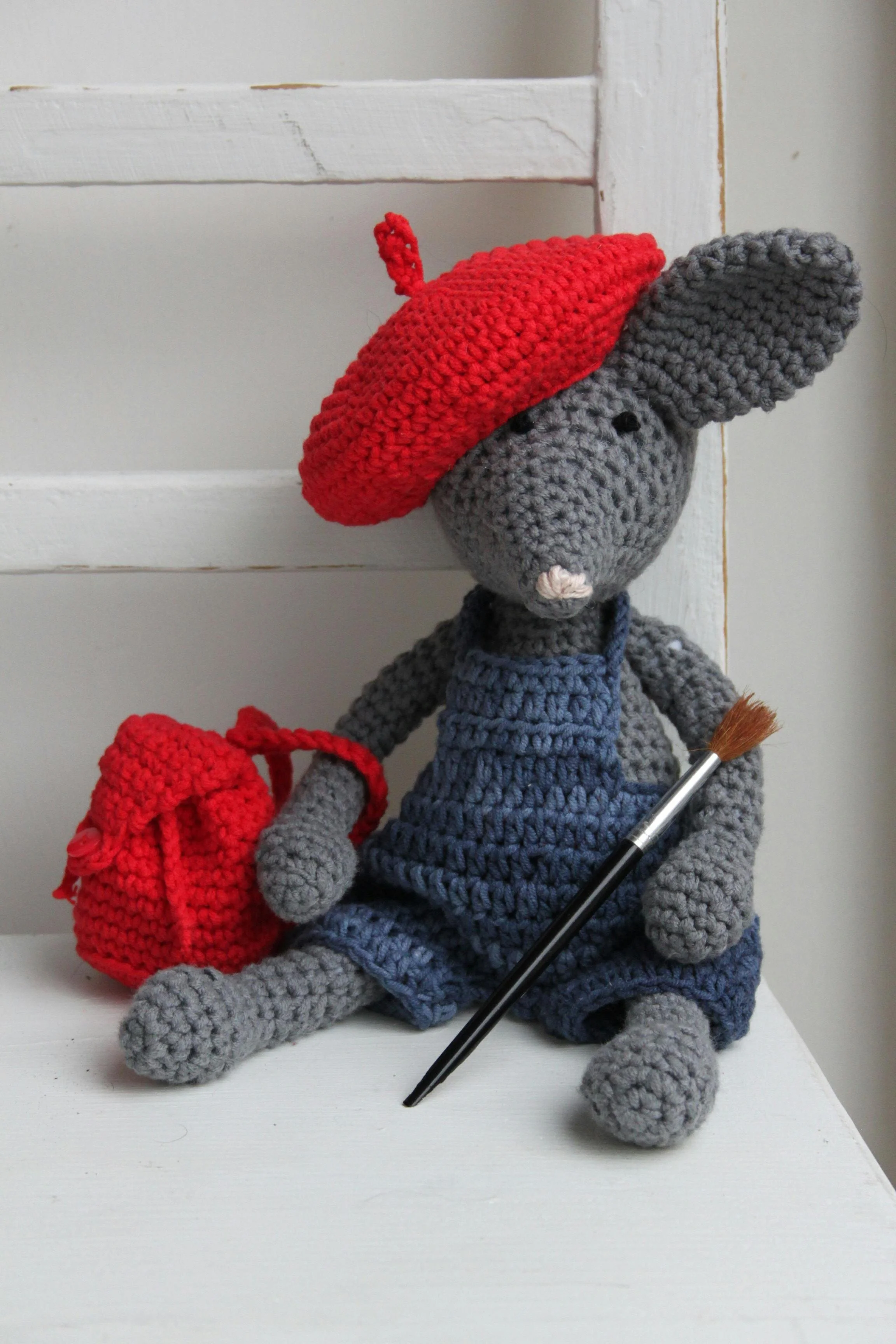 A knitted grey mouse wearing a blue dress and red beret holding a paintbrush and red rucksack