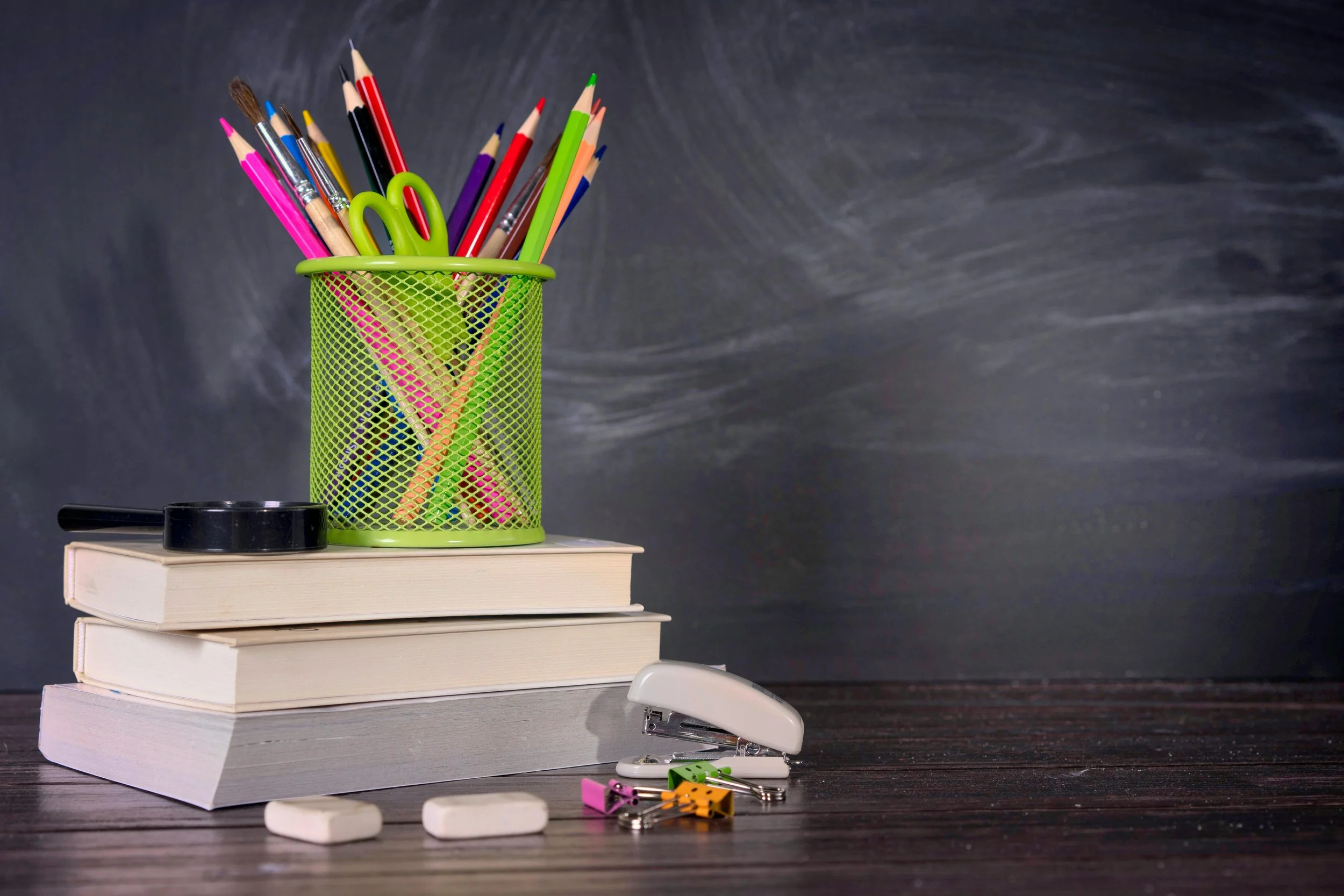 A pile of French tutoring books with a green pot of pencils and scissors on top in front of a blackboard