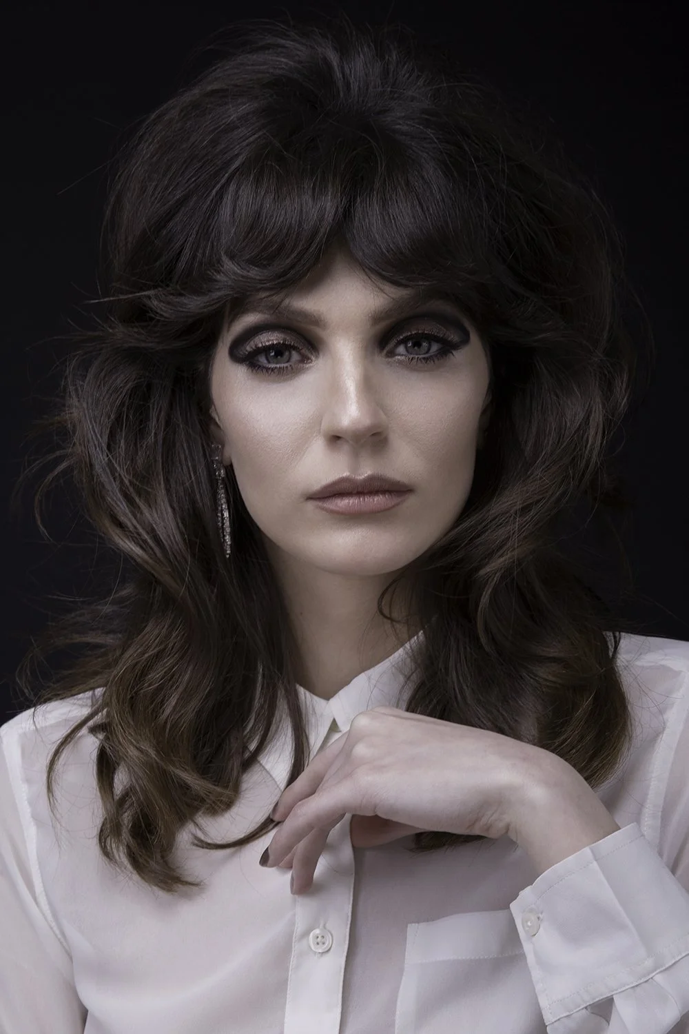 A woman with dark brown, voluminous hair, wearing eye makeup and a white blouse, looking directly at the camera with a neutral expression, against a black background.