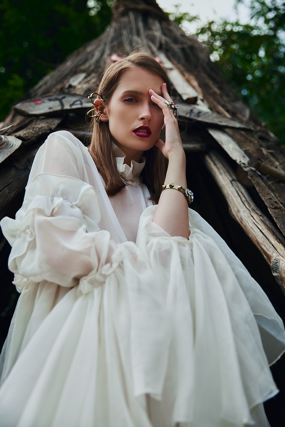 Woman with brown hair and red lipstick posing outdoors in front of a tree, wearing a flowing white dress and jewelry.