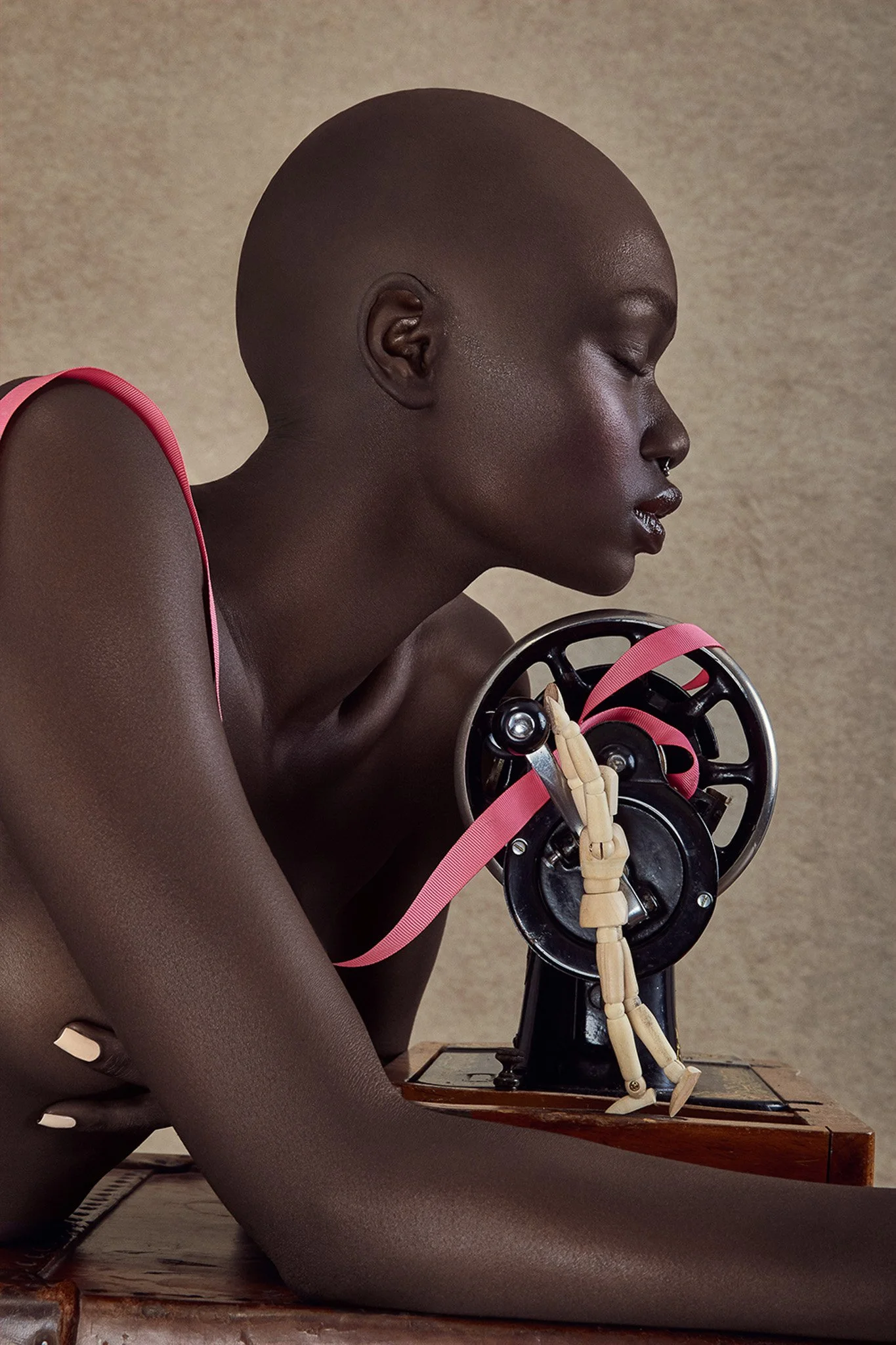 A woman with dark skin and a shaved head holding a mechanical device with a pink ribbon. She is sitting with her eyes closed, and the background is a plain wall.