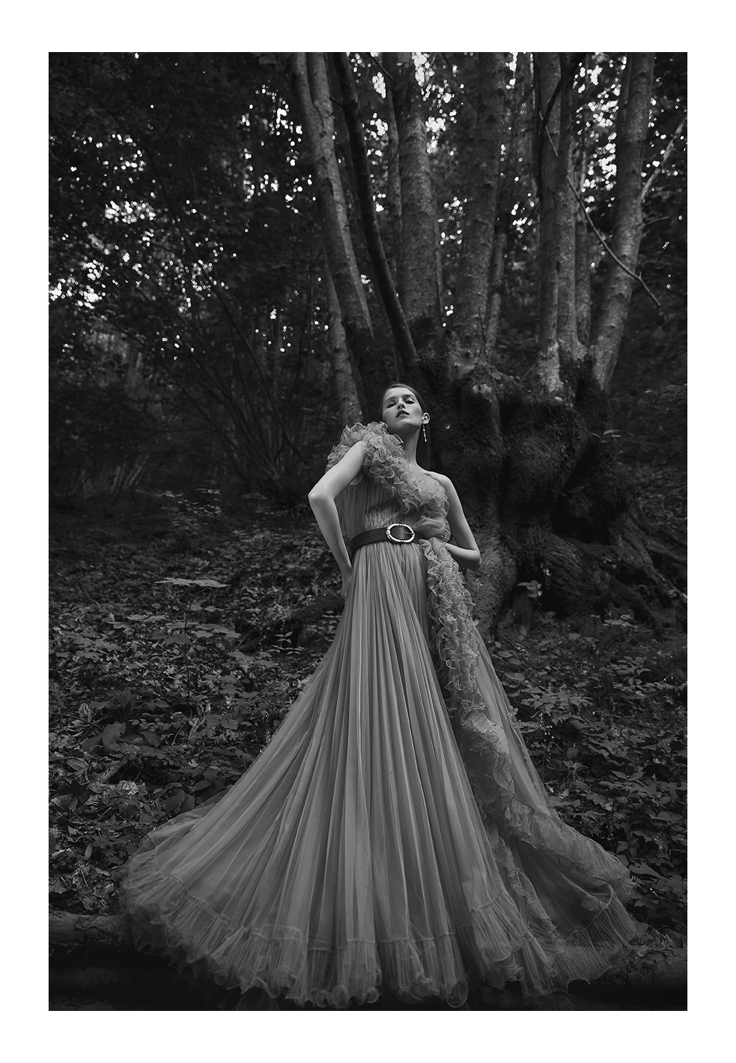 A woman in a flowing, pleated dress with ruffle details poses confidently in a forest setting, black and white photograph