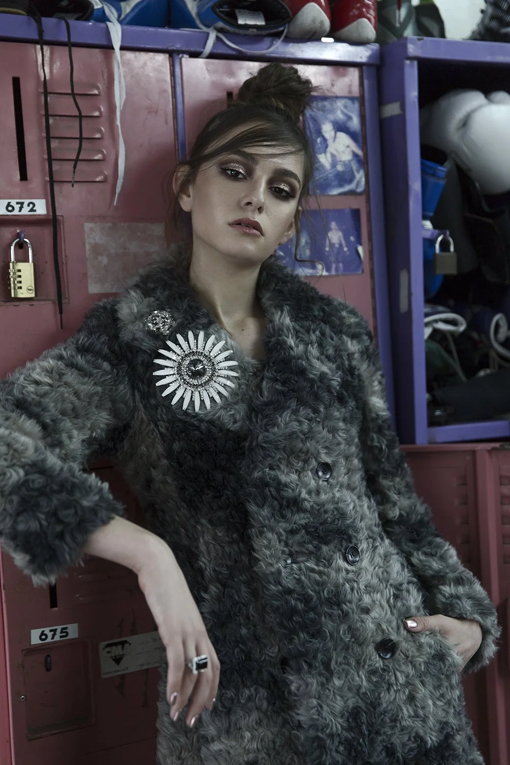 A young woman posing in front of lockers, wearing a curly faux fur coat with decorative pins, showing a confident expression.
