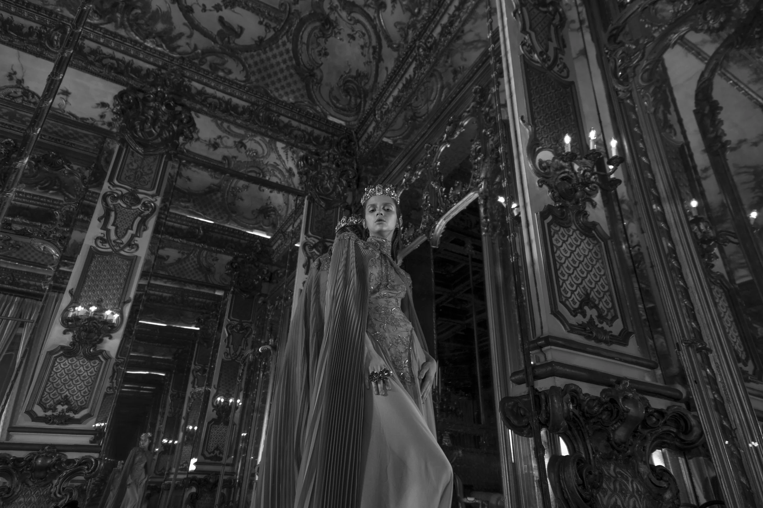 A woman dressed in an ornate gown and wearing a tiara inside an elaborately decorated room with intricate woodwork, gilded accents, and mirrors, surrounded by chandeliers.