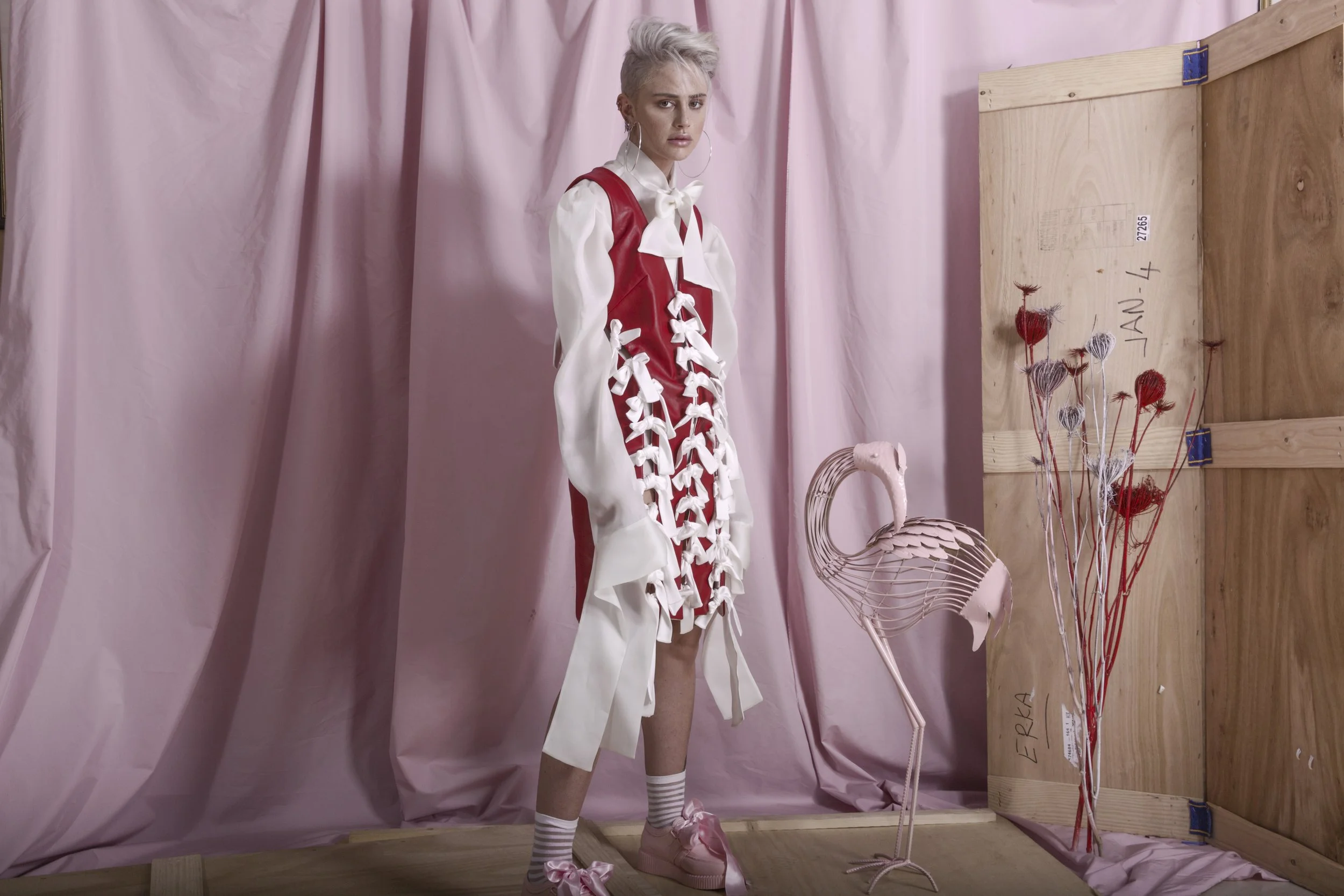 A woman with short platinum blonde hair and hoop earrings stands in front of a pink backdrop. She is wearing a red and white outfit with bows and ribbons, striped socks, and pink shoes. There are various objects next to her, including a pink flamingo