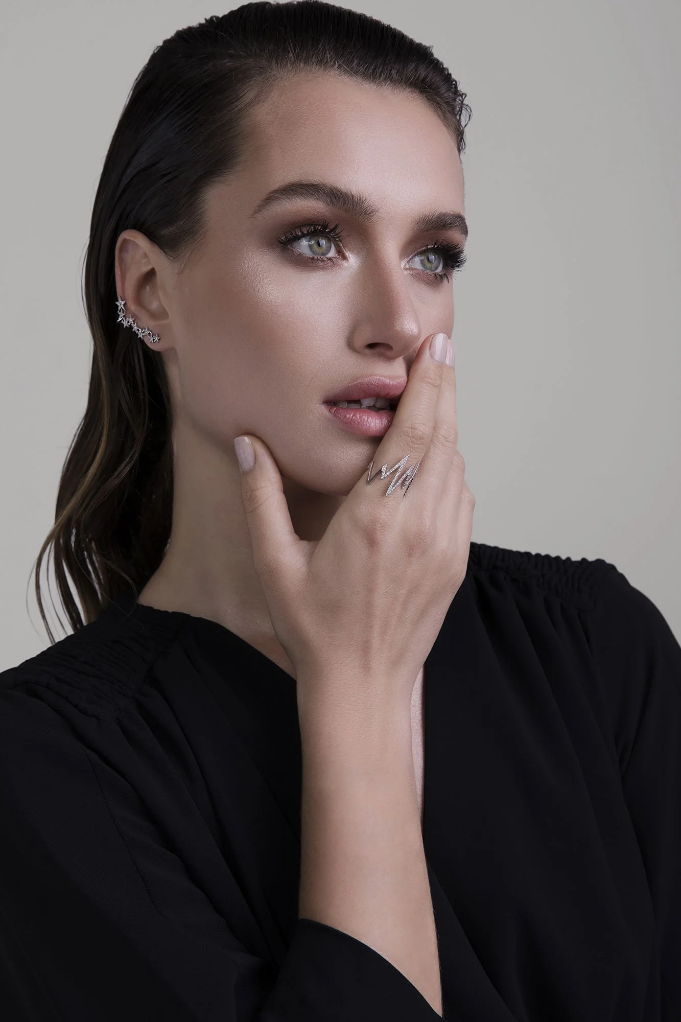 A woman with dark, wet hair and blue eyes poses against a neutral background, touching her face with one hand, wearing a black top, and silver jewelry.