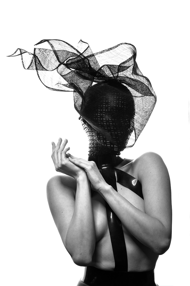 A woman with her face covered by a black mesh veil, no clothing on her upper body, holding her hands close to her chest, with a large black ribbon or strap around her neck and shoulder, in a monochrome photograph.